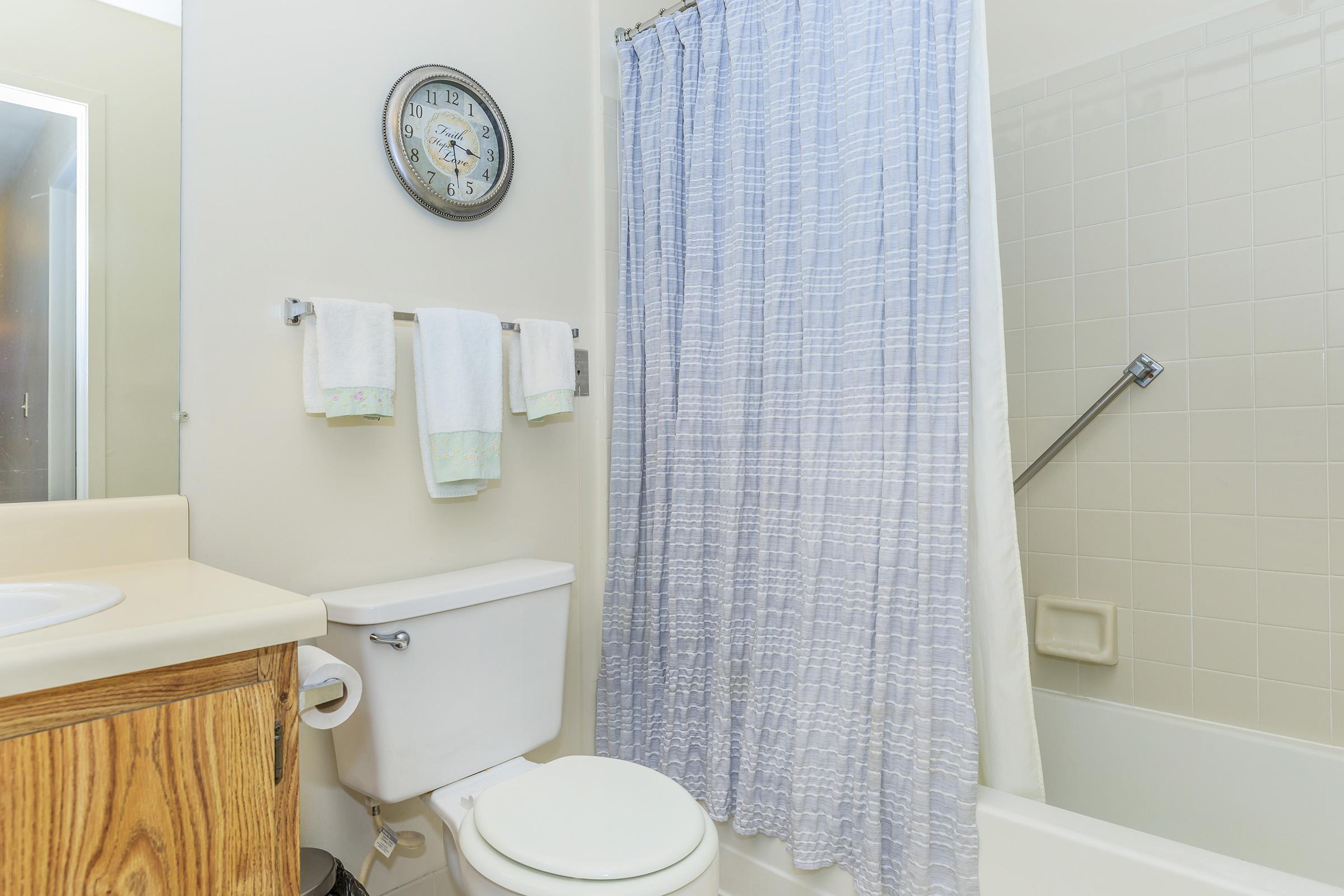 A clean and bright bathroom featuring a bathtub with a shower curtain, a wooden vanity with a sink, a wall clock, and two white towels hanging on a rack. The walls are painted light colors, creating a spacious feel.