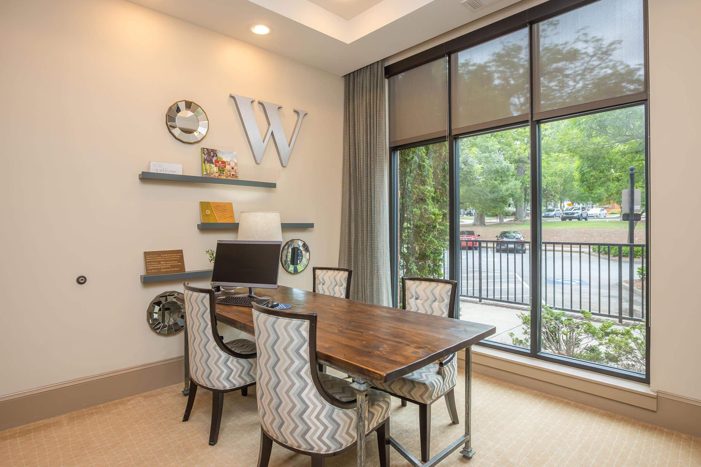 A modern office space featuring a wooden table with chevron-patterned chairs, a computer monitor, and decorative shelves with various items. Large windows allow natural light and provide a view of greenery outside. The walls are painted in neutral tones, enhancing the contemporary aesthetic.