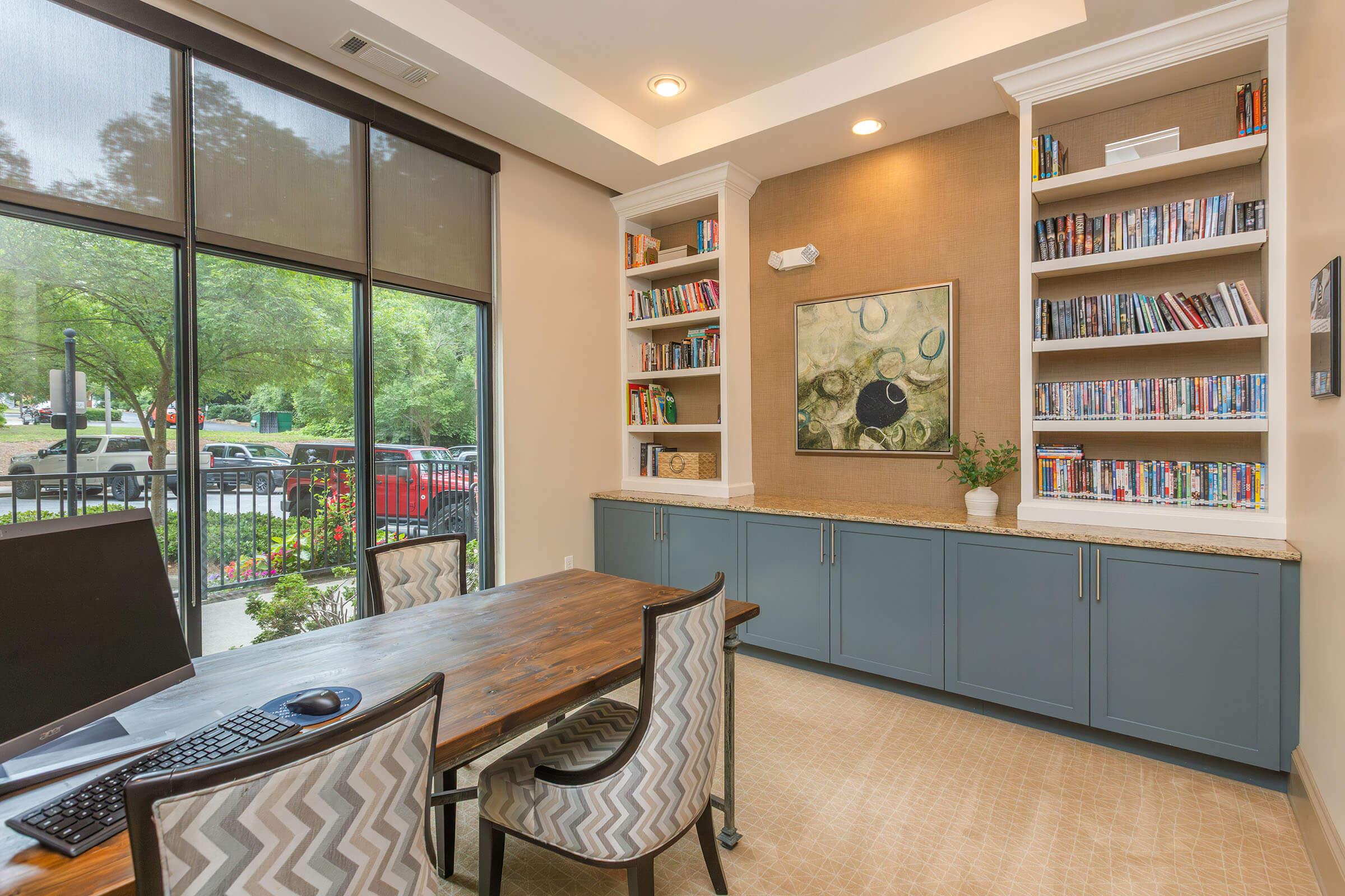 A modern office space featuring a wooden table with two patterned chairs, a computer on the table, and a shelf filled with books. The walls are light-colored, with a framed artwork hanging above a cabinet, and large windows letting in natural light, overlooking greenery outside.