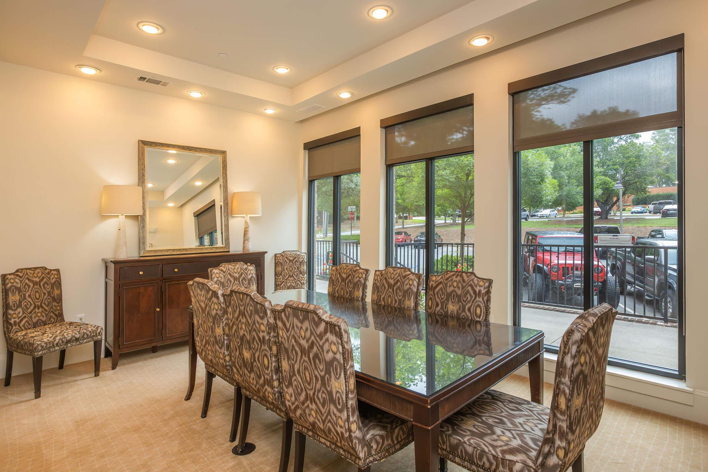 A spacious dining area featuring a large glass table surrounded by patterned chairs, with a wooden sideboard against the wall. Large windows allow natural light to fill the room, offering a view of trees and parked cars outside. Brightly lit with modern ceiling lights and a mirror on the wall.