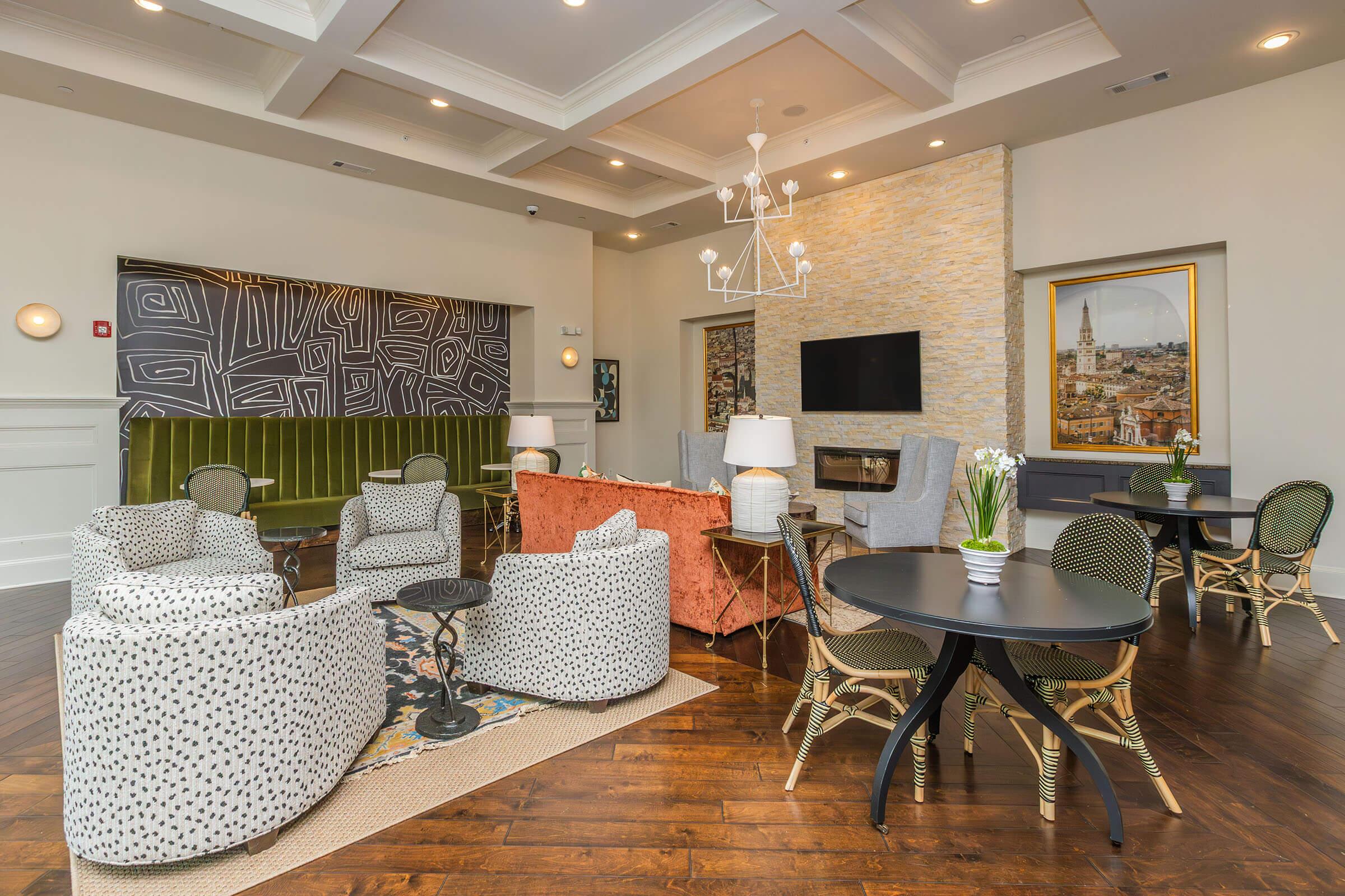 A stylish lounge area with a mix of patterned seating, including two polka-dot chairs, an orange couch, and round tables. The space features a neutral color palette, modern light fixtures, and a stone accent wall with a TV. Decorative elements like green plants and artwork add a touch of elegance.