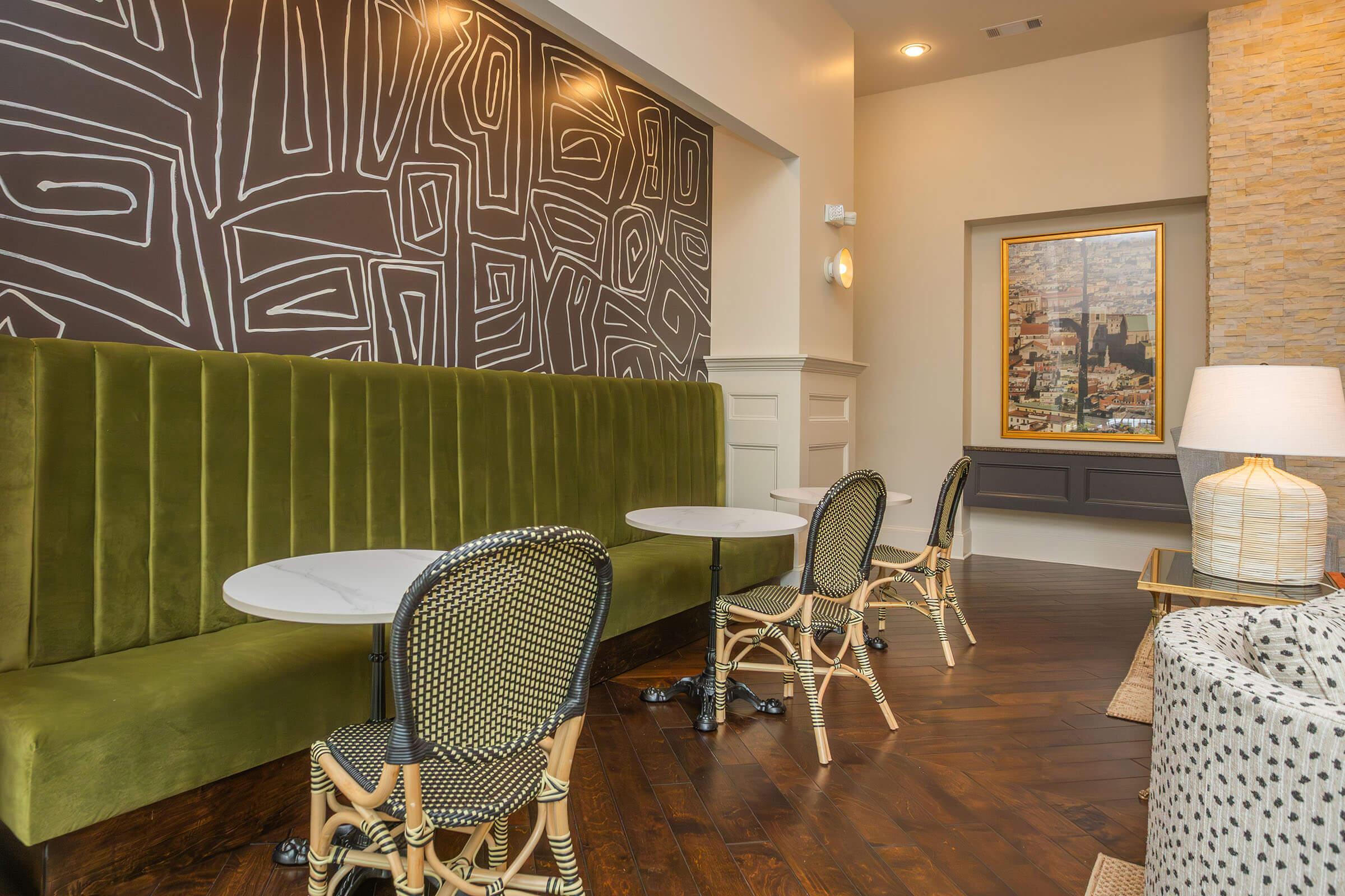 A cozy interior featuring a green velvet bench along the wall, small round tables in front, and stylish woven chairs. The background has a modern abstract design and a framed painting of a cityscape, with warm lighting enhancing the inviting atmosphere.