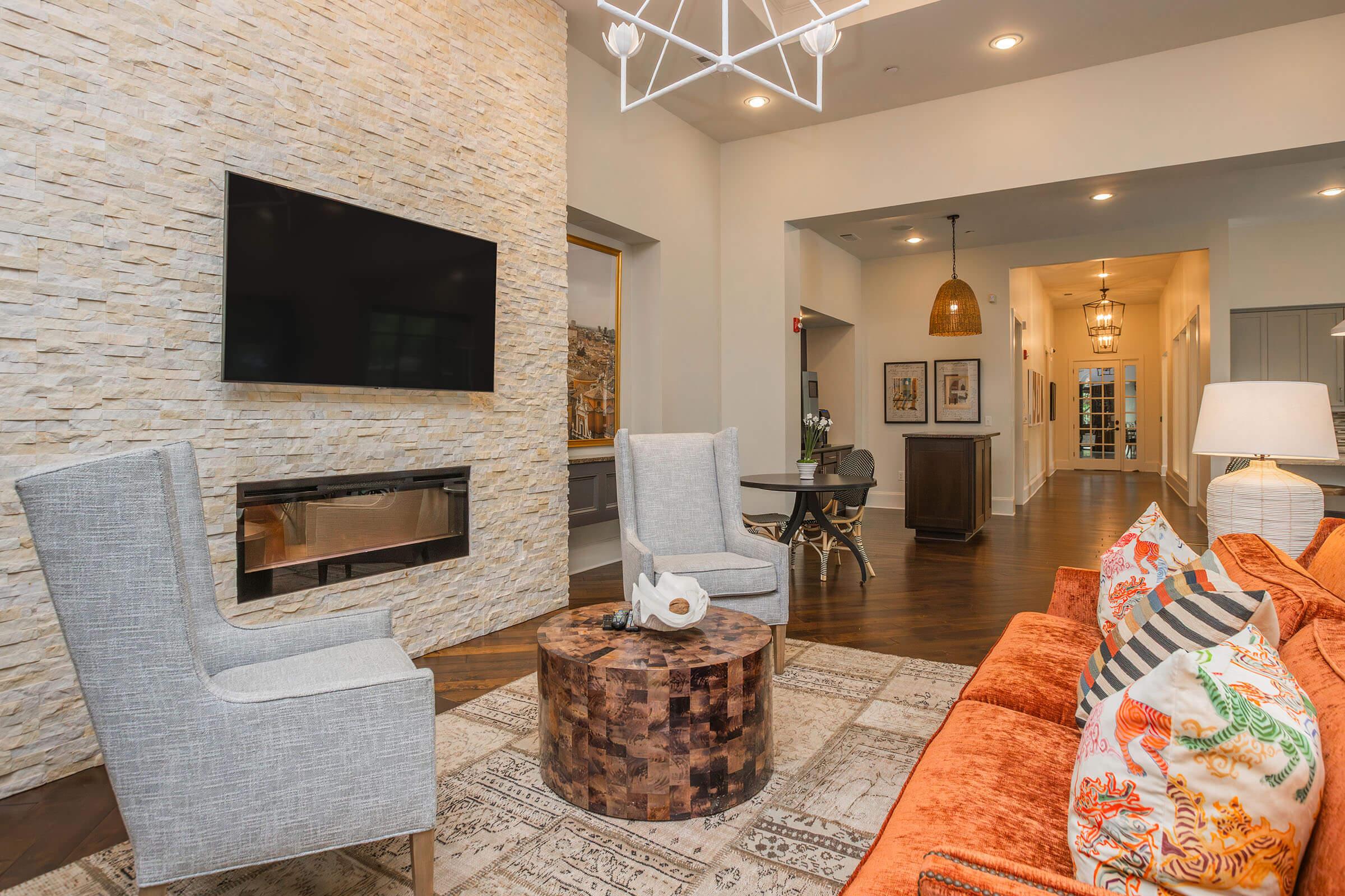 A modern living room with a stone accent wall, featuring two light gray armchairs and an orange sofa decorated with colorful cushions. A circular wooden coffee table sits in the center, and a flat-screen TV is mounted on the wall above an electric fireplace. The space is well-lit with stylish light fixtures.