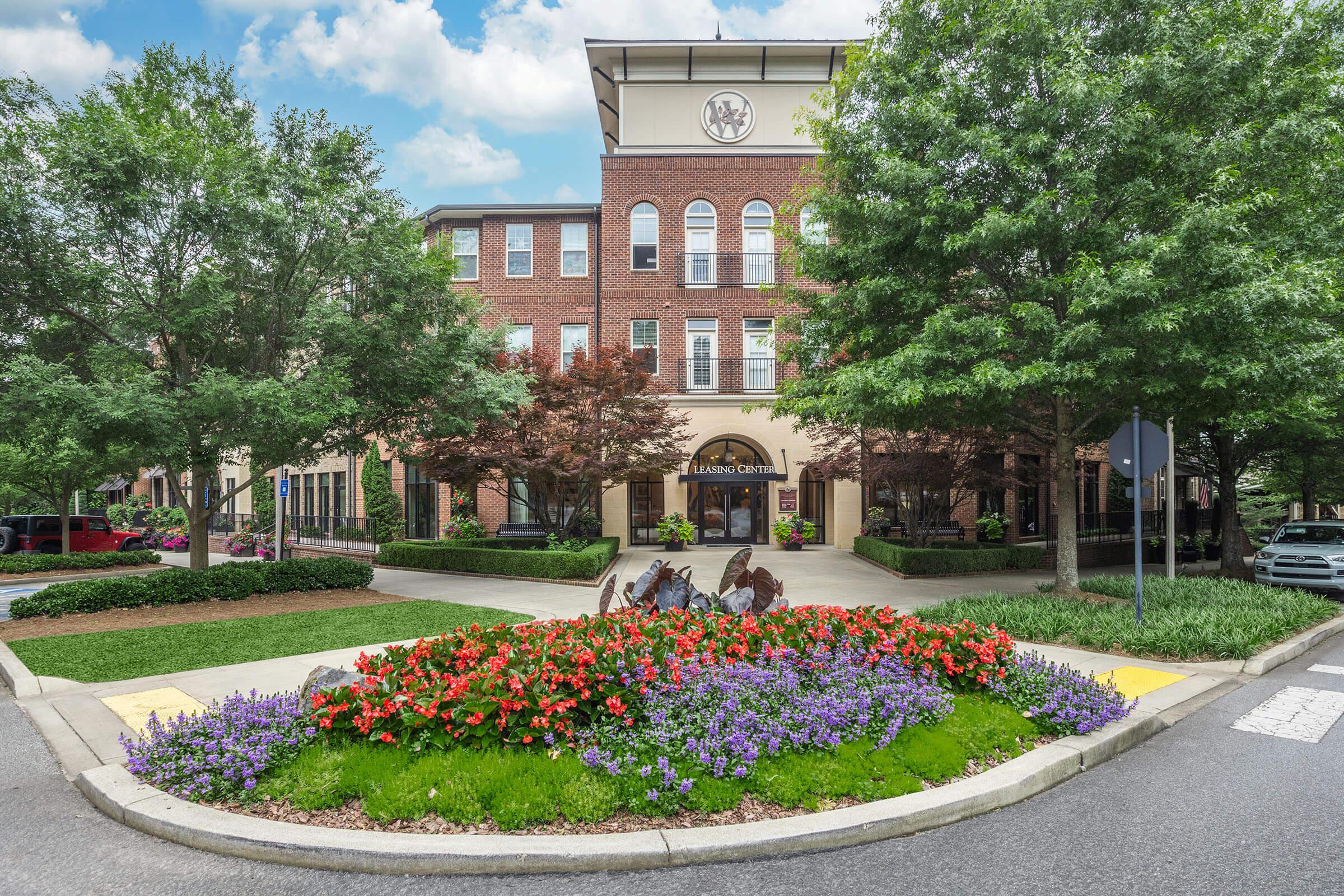 A landscaped roundabout featuring colorful flower beds with red and purple blooms. In the background, a brick building with large windows and an elegant entrance is visible, surrounded by trees. The scene is bright and inviting, suggesting a well-maintained area in an urban setting.
