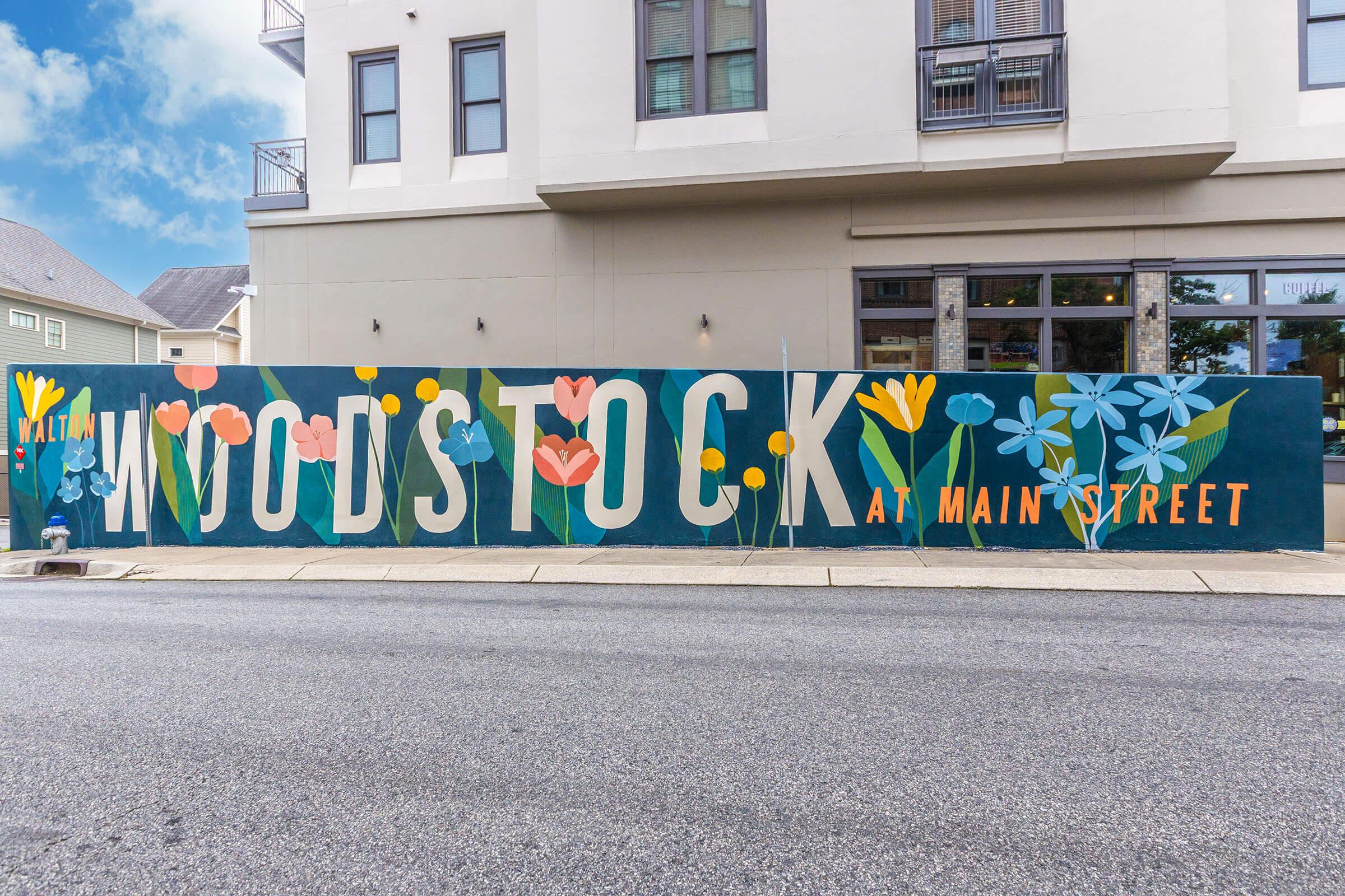 A colorful mural on a building featuring the text "WALTON WOODSTOCK AT MAIN STREET" in large letters, surrounded by vibrant flowers and greenery. The mural adds a cheerful touch to the urban setting, with a clear blue sky and a street view in the foreground.