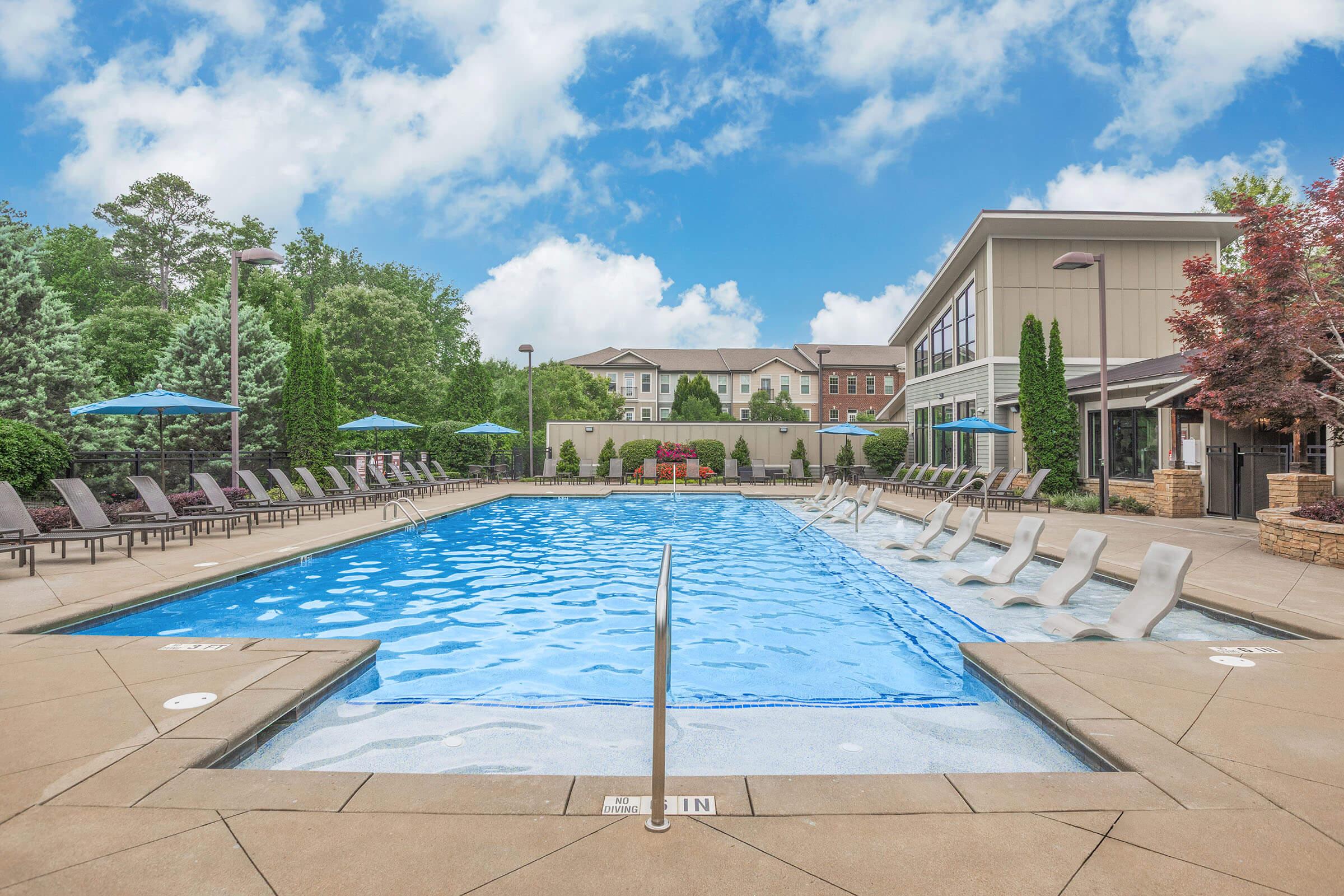 A large outdoor swimming pool surrounded by lounge chairs and umbrellas, with trees and buildings in the background. The sky is partly cloudy, creating a bright and inviting atmosphere. The pool features a gentle slope leading into the water, making it accessible for all.