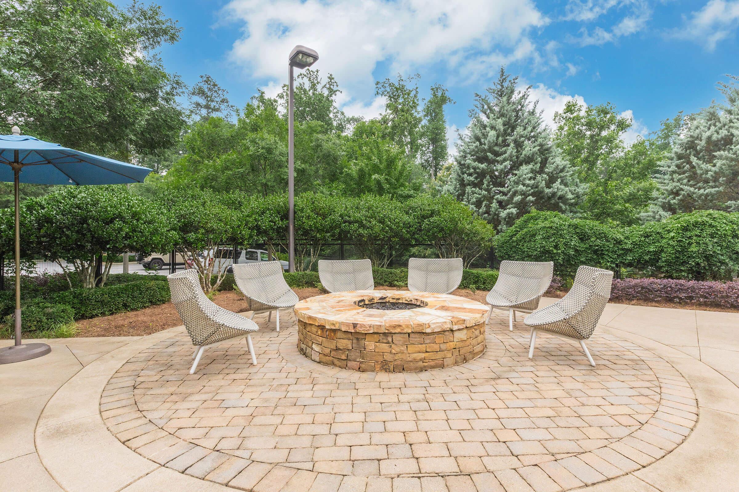Outdoor seating area featuring a stone fire pit surrounded by six modern chairs on a circular paved patio. Lush greenery and trees provide a natural backdrop, with a blue umbrella offering shade. The sky is partly cloudy, creating a serene and inviting atmosphere.