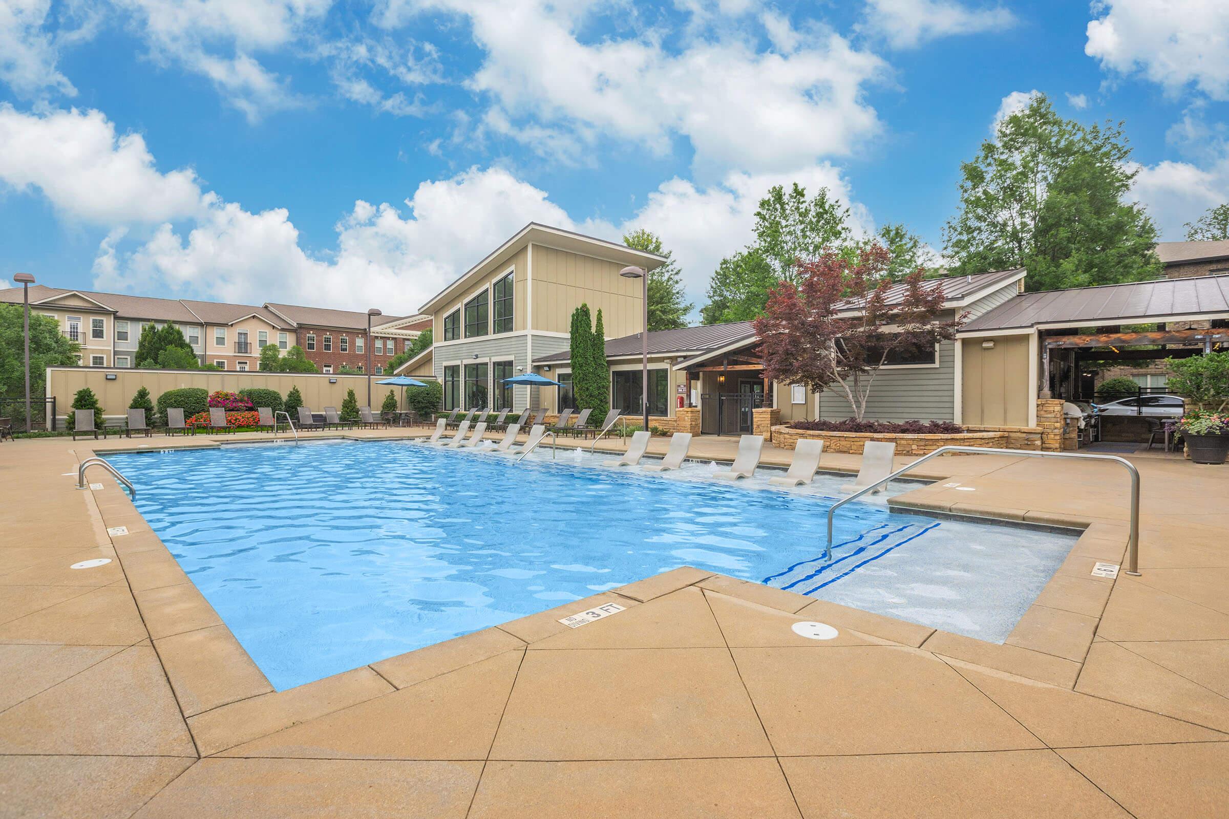 Outdoor swimming pool surrounded by a patio with lounge chairs. A modern building is visible in the background, featuring large windows and green landscaping. The sky is partly cloudy, creating a serene atmosphere.