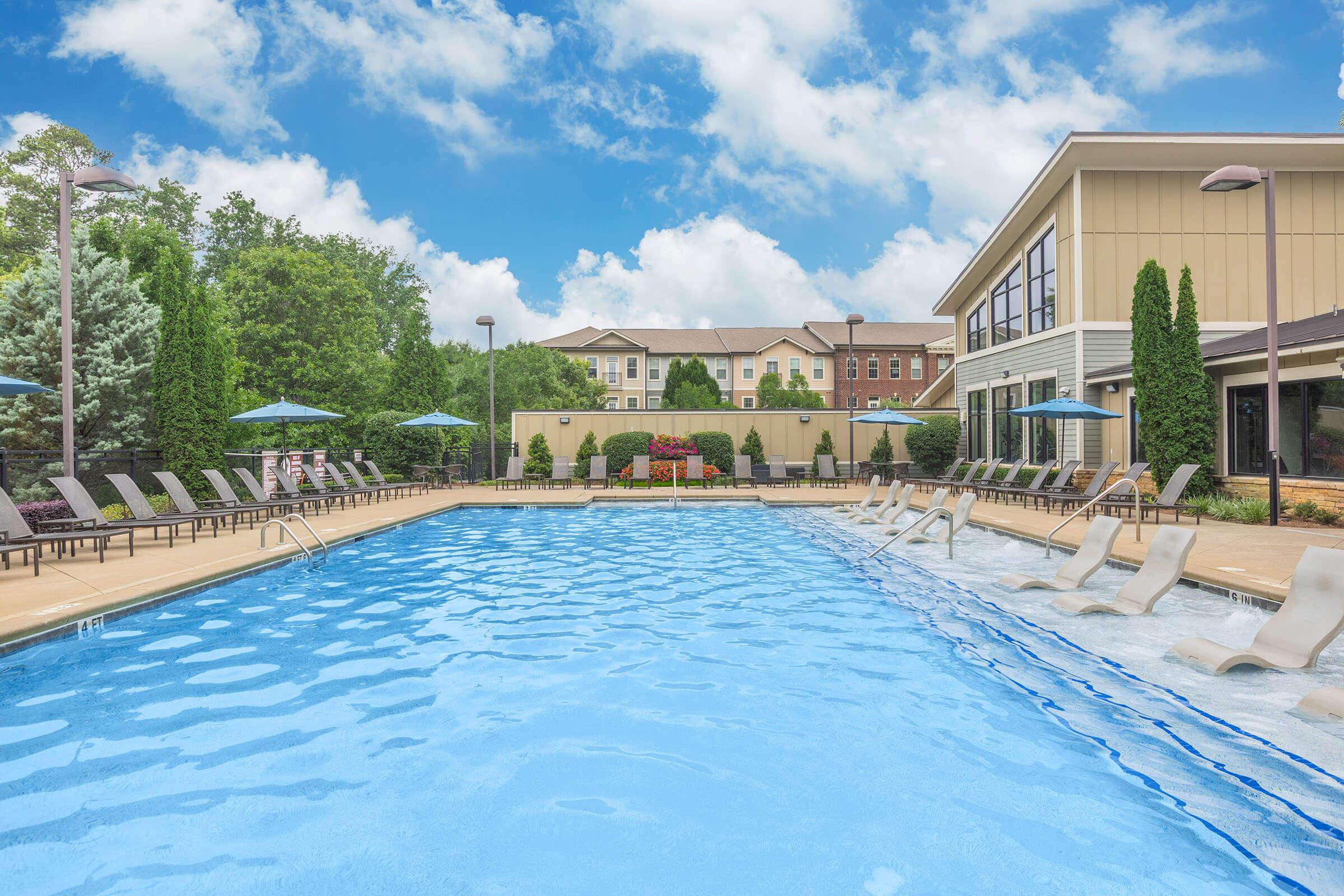 A clear blue swimming pool surrounded by lounge chairs and umbrellas. Lush green trees line the area, and a modern building is visible in the background under a bright, partly cloudy sky. The pool area is inviting and well-maintained, ideal for relaxation and leisure activities.