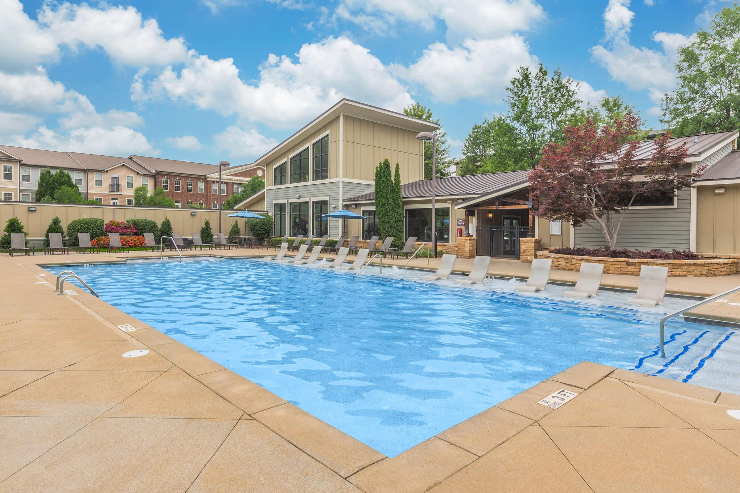 A large, clear swimming pool surrounded by lounge chairs, with well-maintained landscaping and a modern building in the background. The sky is partly cloudy, adding a bright and inviting atmosphere to the area.
