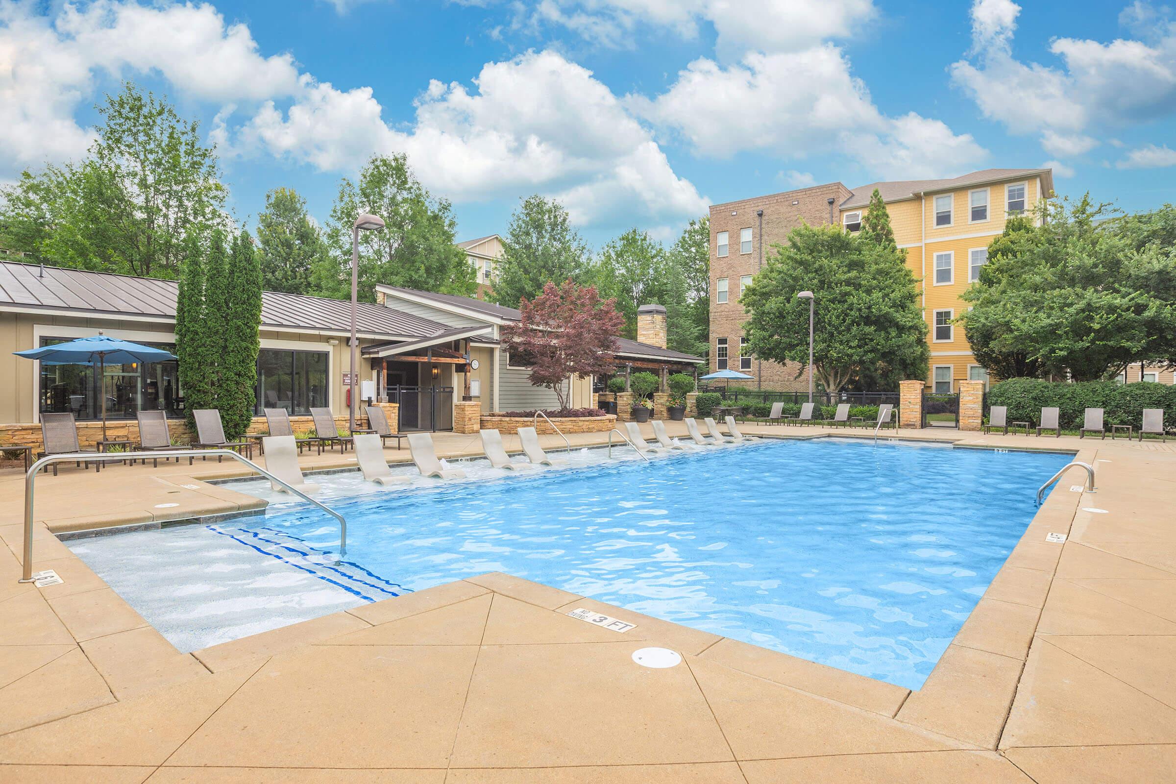 A large outdoor swimming pool with lounge chairs arranged around the perimeter. Surrounding the pool are green trees and shrubs, with a stylish building in the background. The sky is partly cloudy, creating a bright and inviting atmosphere. The scene conveys a relaxing, resort-like vibe.