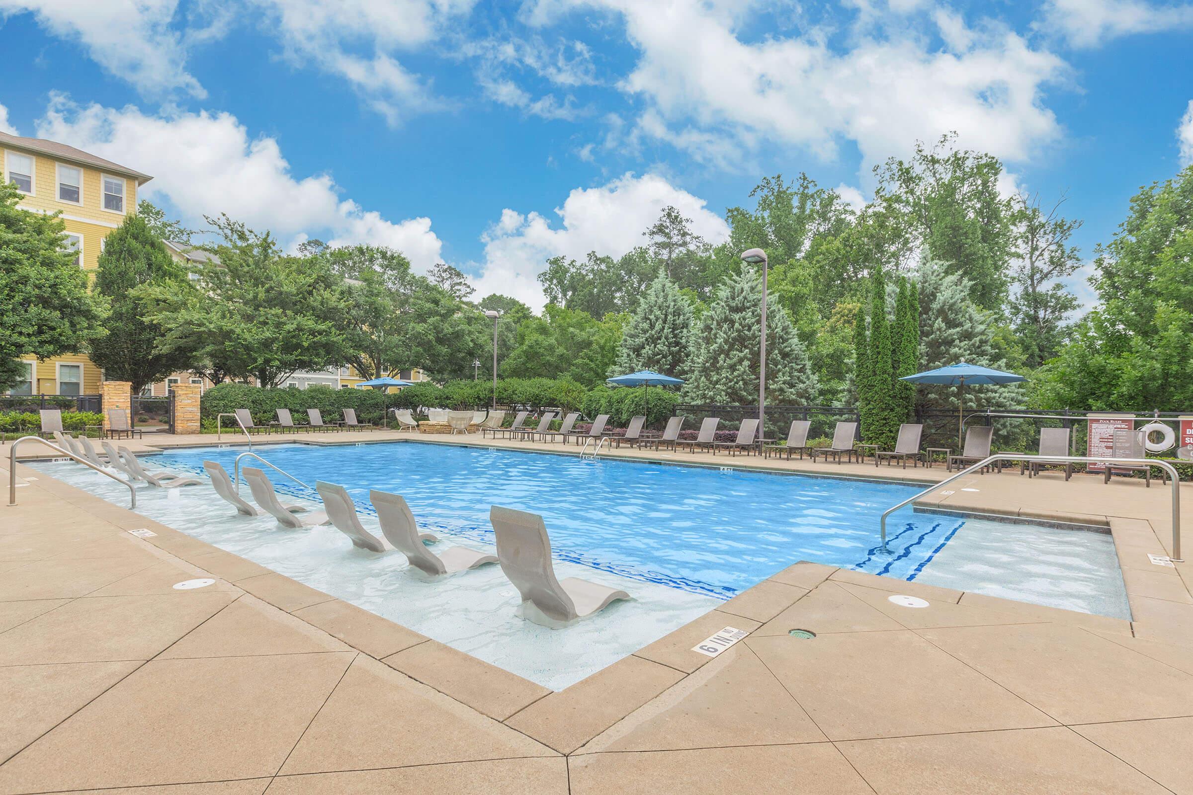 A clear blue swimming pool surrounded by lounge chairs and umbrellas, set in a landscaped area with trees and greenery. The sky is bright with clouds, creating a serene atmosphere.
