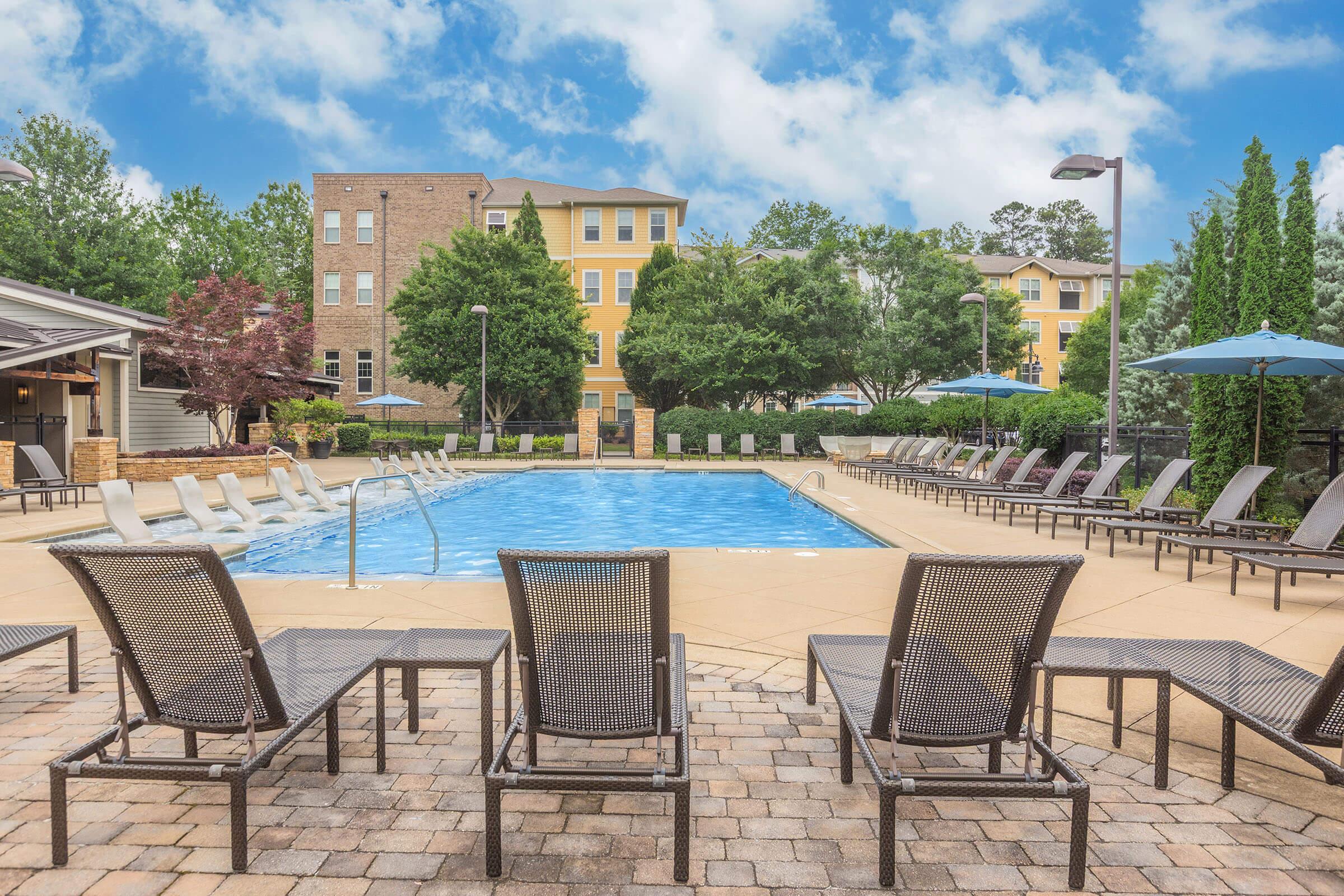 A serene outdoor pool area featuring a clear blue pool surrounded by lounge chairs and umbrellas. Lush trees and greenery provide a relaxing atmosphere, while modern apartment buildings are visible in the background under a cloudy sky.