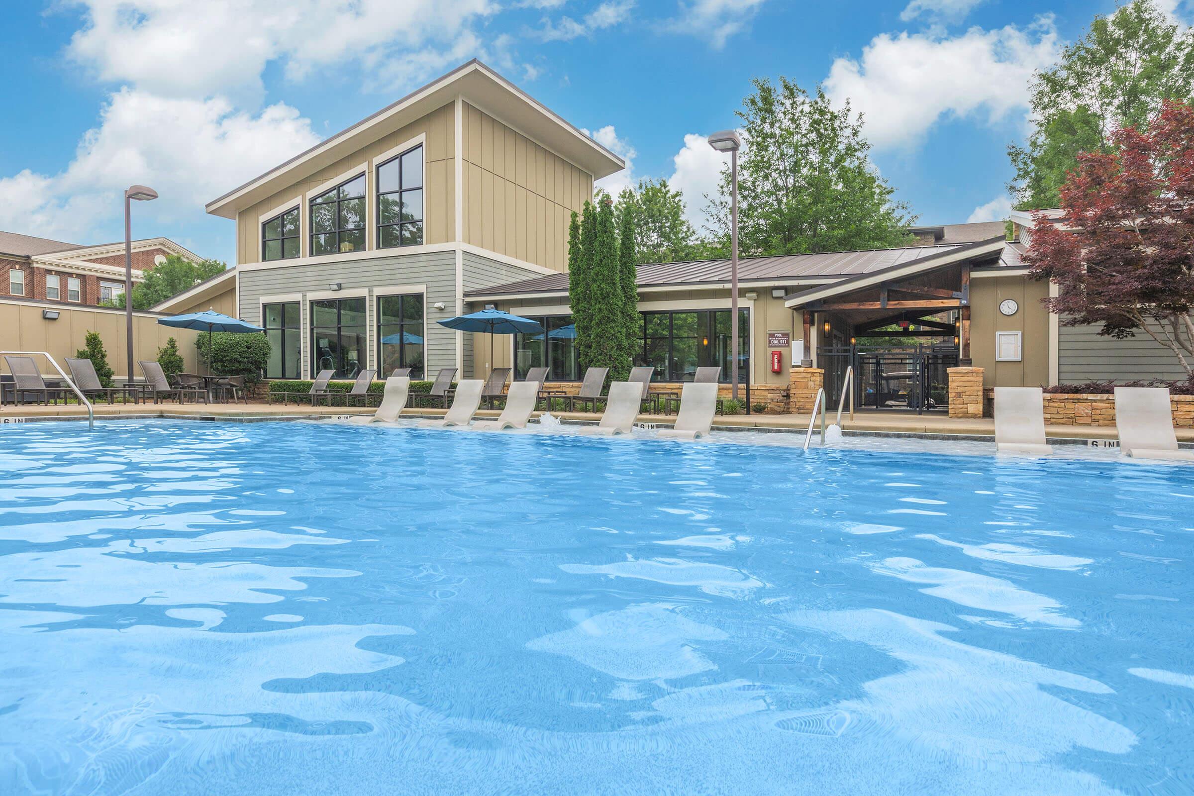 A clear blue swimming pool surrounded by lounge chairs, with a modern building in the background featuring large windows and outdoor seating. Lush green trees and shrubs enhance the serene atmosphere under a bright blue sky with scattered clouds.