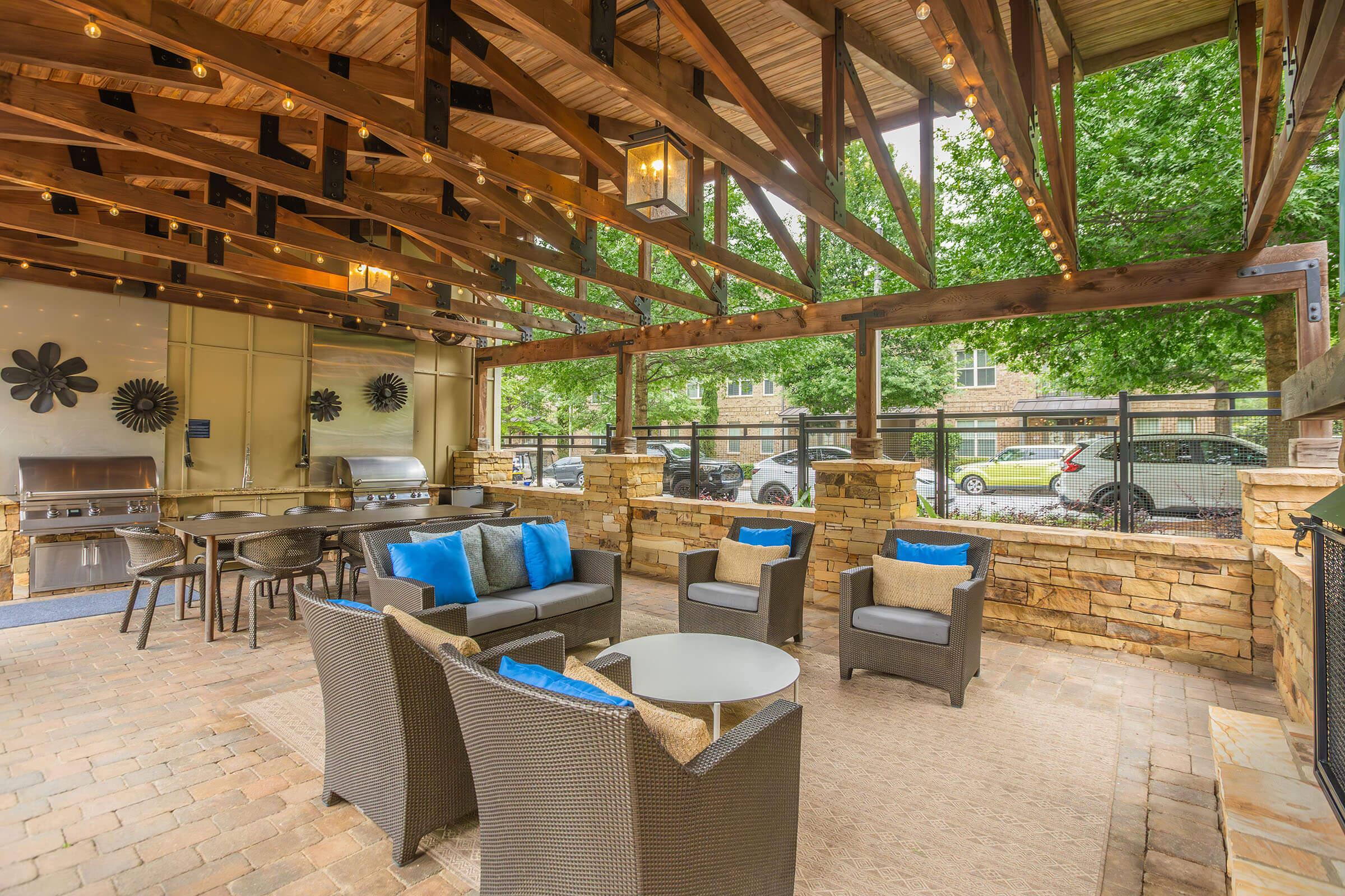 An outdoor patio area featuring a wooden pergola, comfortable seating with blue accent pillows, and a glass-top table. The space is well-lit with hanging lights, surrounded by greenery, and includes a stone wall. Grill equipment can be seen in the background, creating a relaxed atmosphere for gatherings.