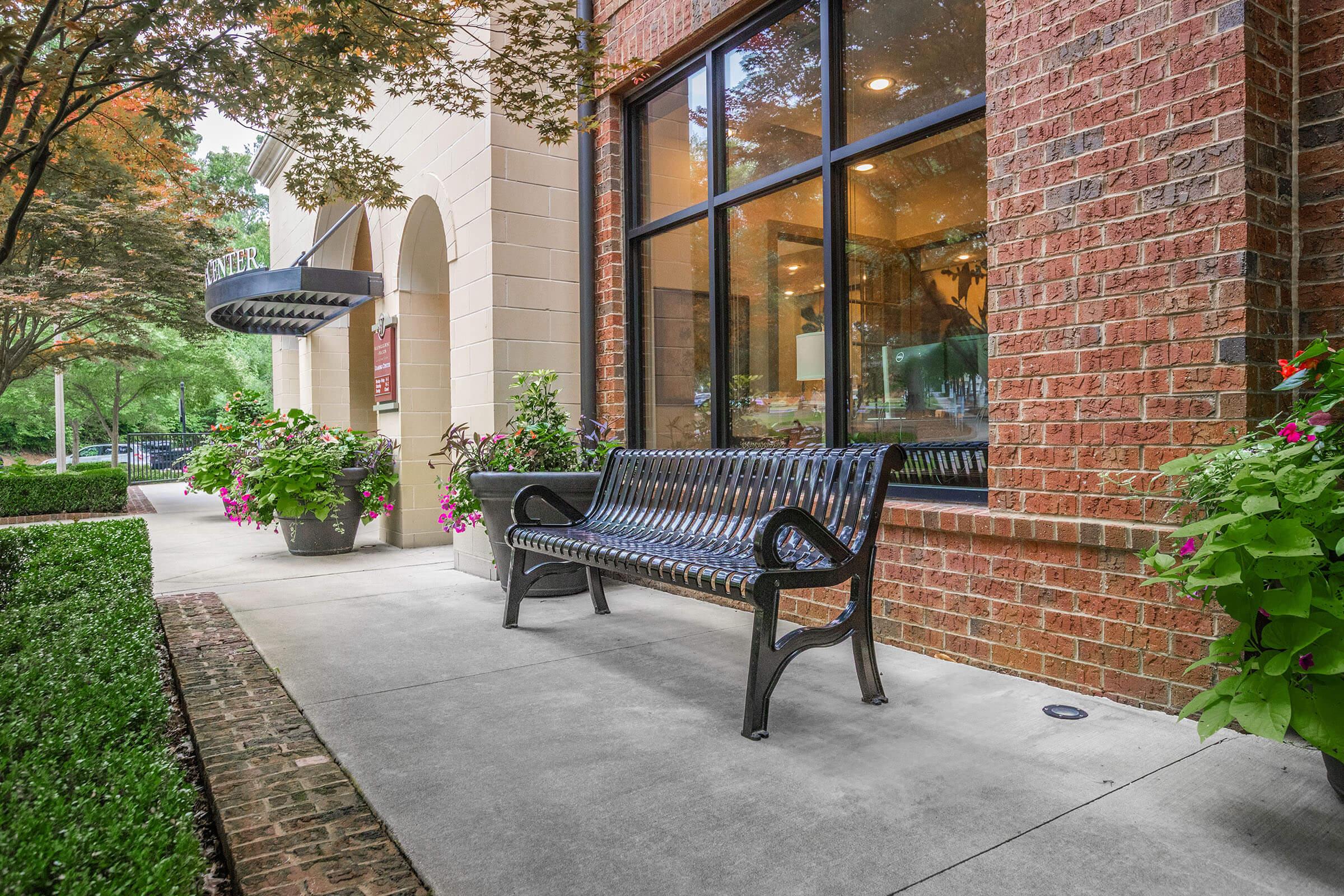 A black metal bench sits on a concrete walkway in front of a brick building with large windows. Colorful flower pots are placed around the bench, and neatly trimmed hedges line the pathway. The scene exudes a welcoming atmosphere with greenery and decorative plants.