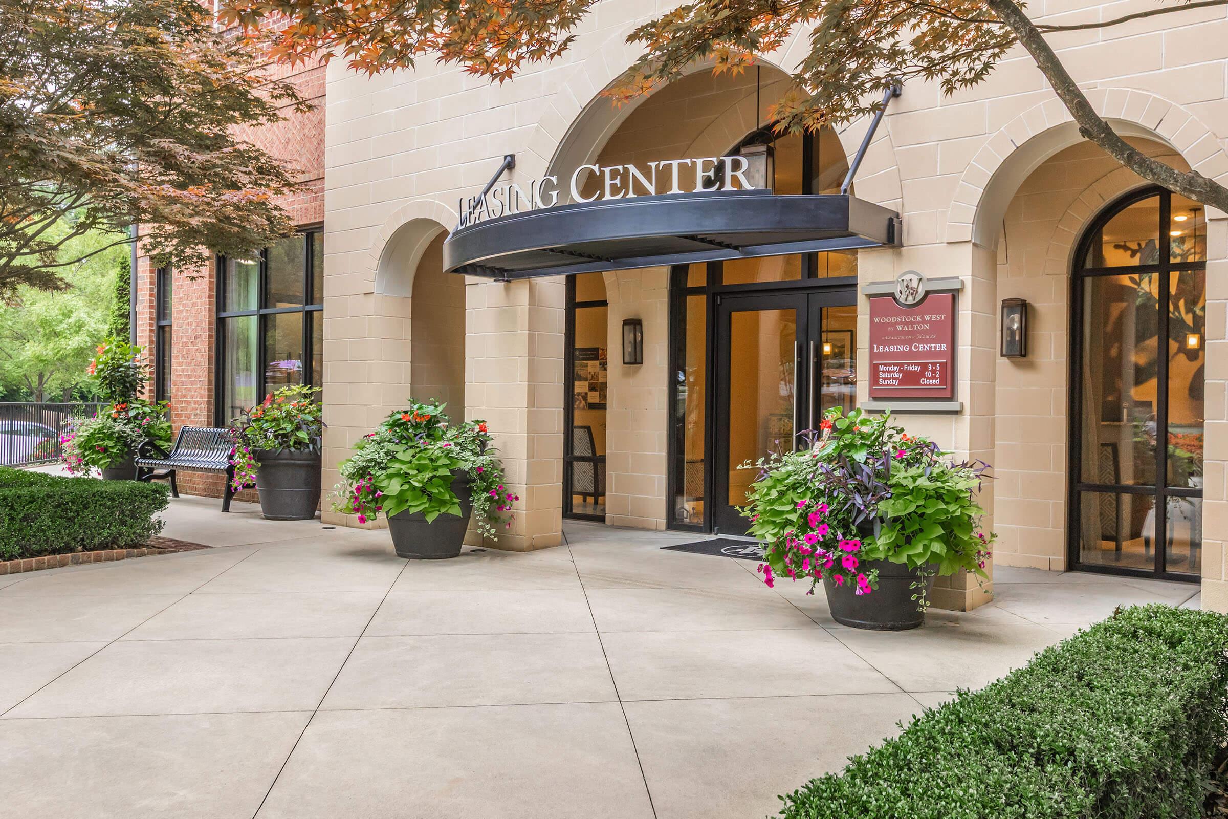 Entrance to a building labeled "Lansing Center," featuring a black awning and decorative planters filled with vibrant flowers. The pathway is paved and bordered by neatly trimmed shrubs. Benches are placed nearby, suggesting a welcoming atmosphere.