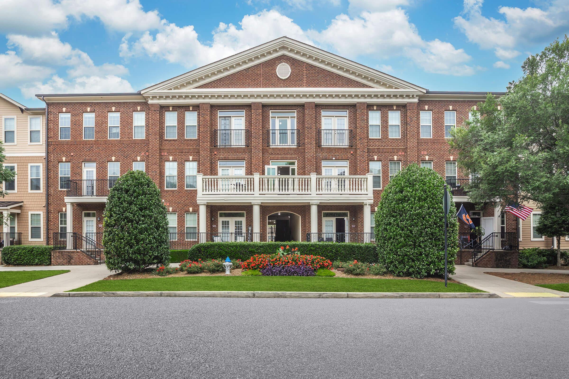 A brick apartment building with a symmetrical design, featuring multiple balconies on the second floor. Flanked by neatly trimmed hedges and colorful flower beds in front, the structure is set against a backdrop of blue sky with scattered clouds. An American flag is visible on the right side.