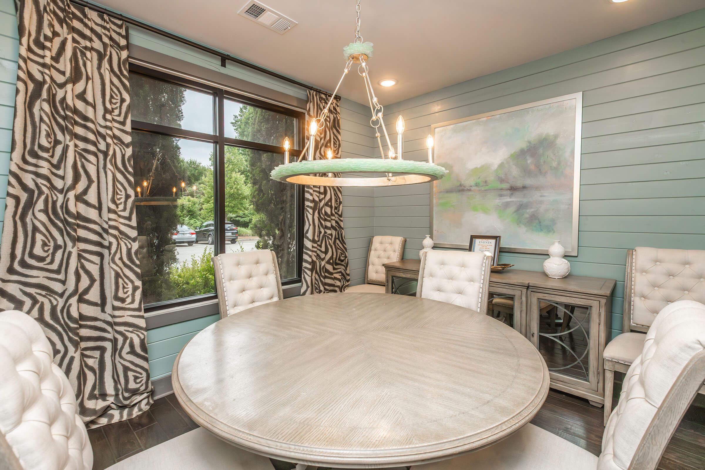 A modern dining room featuring a round wooden table surrounded by upholstered chairs. The walls are painted in a light blue hue with decorative wall panels. Large windows with patterned curtains let in natural light, and a contemporary chandelier hangs above the table. A piece of abstract art is displayed on the wall.