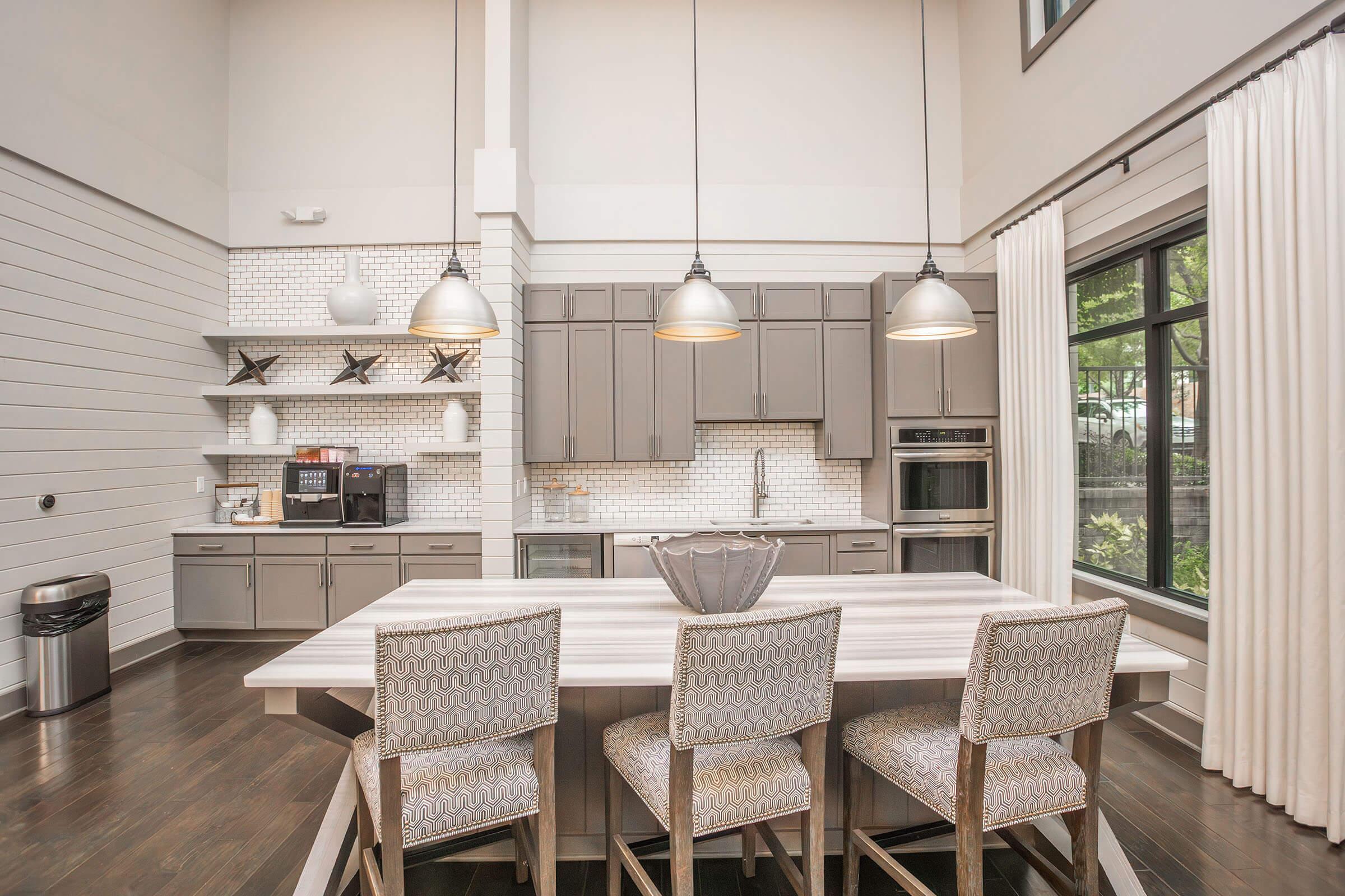Modern kitchen featuring gray cabinetry, a large island with seating for four, pendant lighting, and stainless steel appliances. A window offers natural light with a view of greenery outside. The decor includes minimalist shelves and stylish curtains, creating a contemporary, inviting atmosphere.