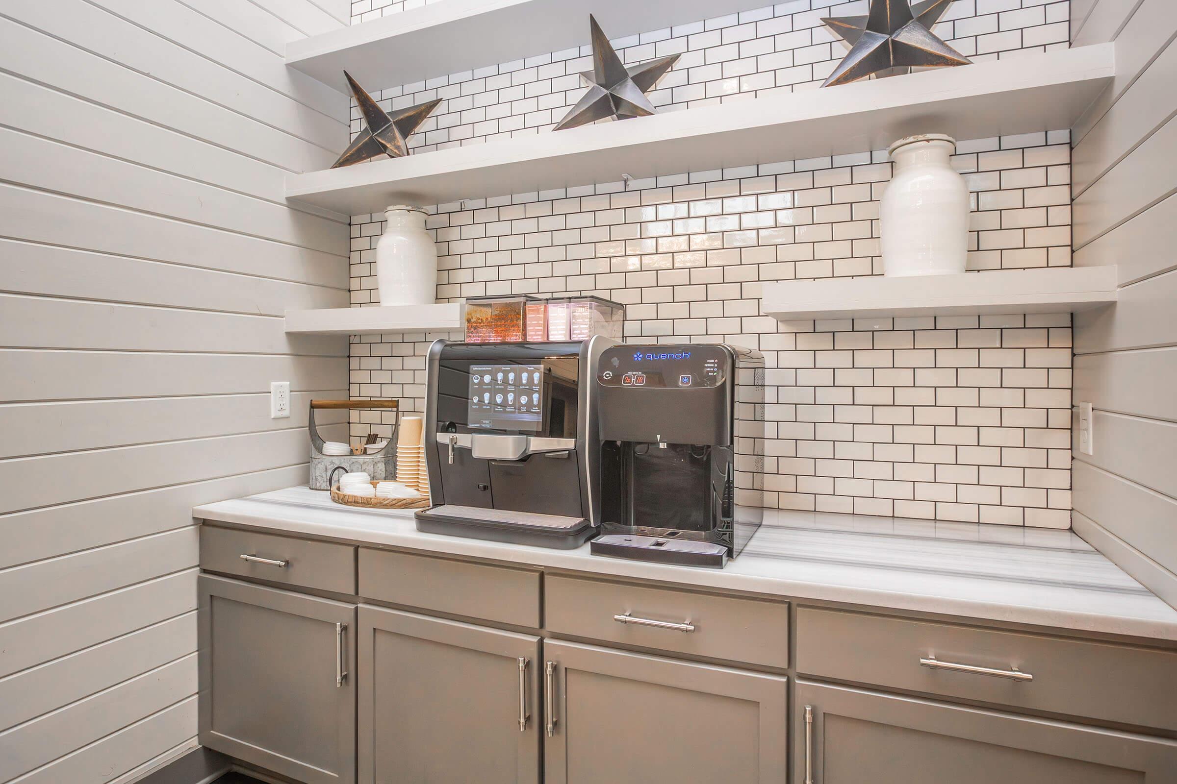 A modern kitchenette featuring two coffee machines on a sleek countertop. Above, decorative shelves hold white vases and star-shaped decor against a tiled backsplash. The cabinetry is light gray, contributing to a clean and contemporary aesthetic.