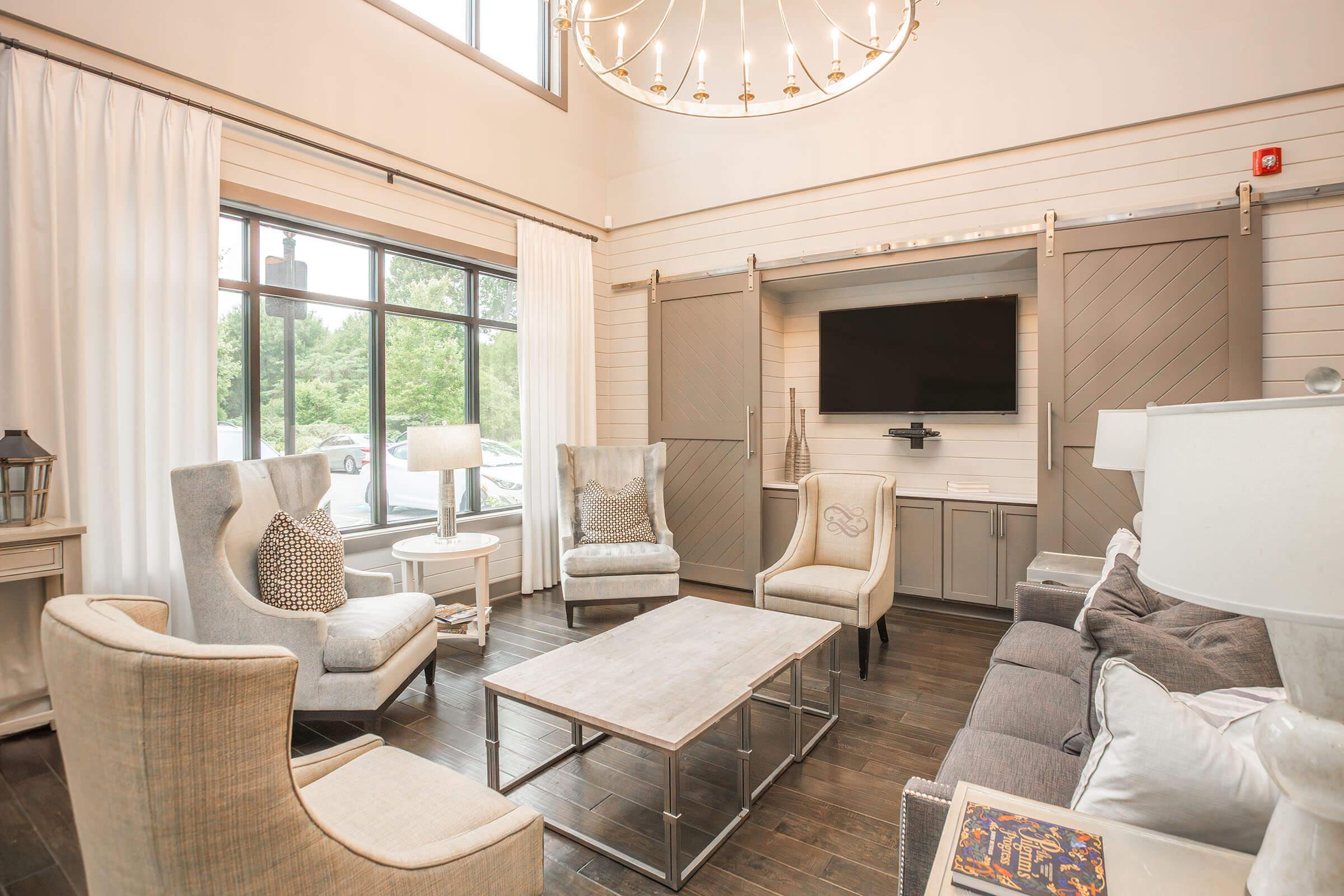 A cozy living area featuring two beige armchairs and a gray sofa. A light wooden coffee table is placed in the center. Large windows with white drapes provide natural light, and a modern chandelier hangs from the ceiling. A TV is mounted on the wall, enhancing the inviting atmosphere of the space.