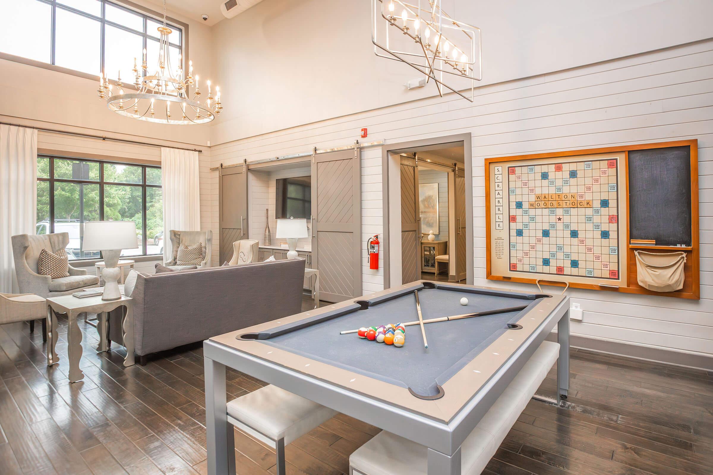 A spacious room featuring a pool table in the foreground, with colorful billiard balls resting on it. In the background, there’s a cozy seating area with elegant chairs and a large window allowing natural light. A decorative Scrabble board is mounted on the wall, enhancing the inviting atmosphere.