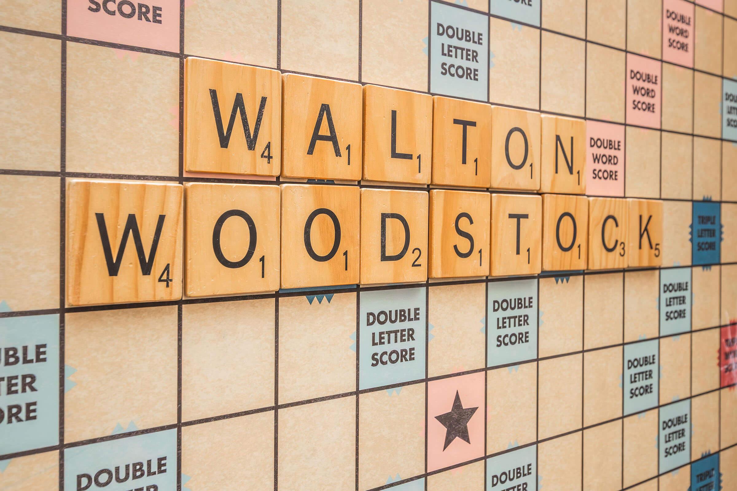 A close-up view of a Scrabble board with wooden letter tiles arranged to spell "WALTON" and "WOODSTOCK." The tiles are positioned on a grid featuring the game's scoring zones, including double word and double letter score spaces. The background shows a light-colored, patterned surface typical of a Scrabble board.