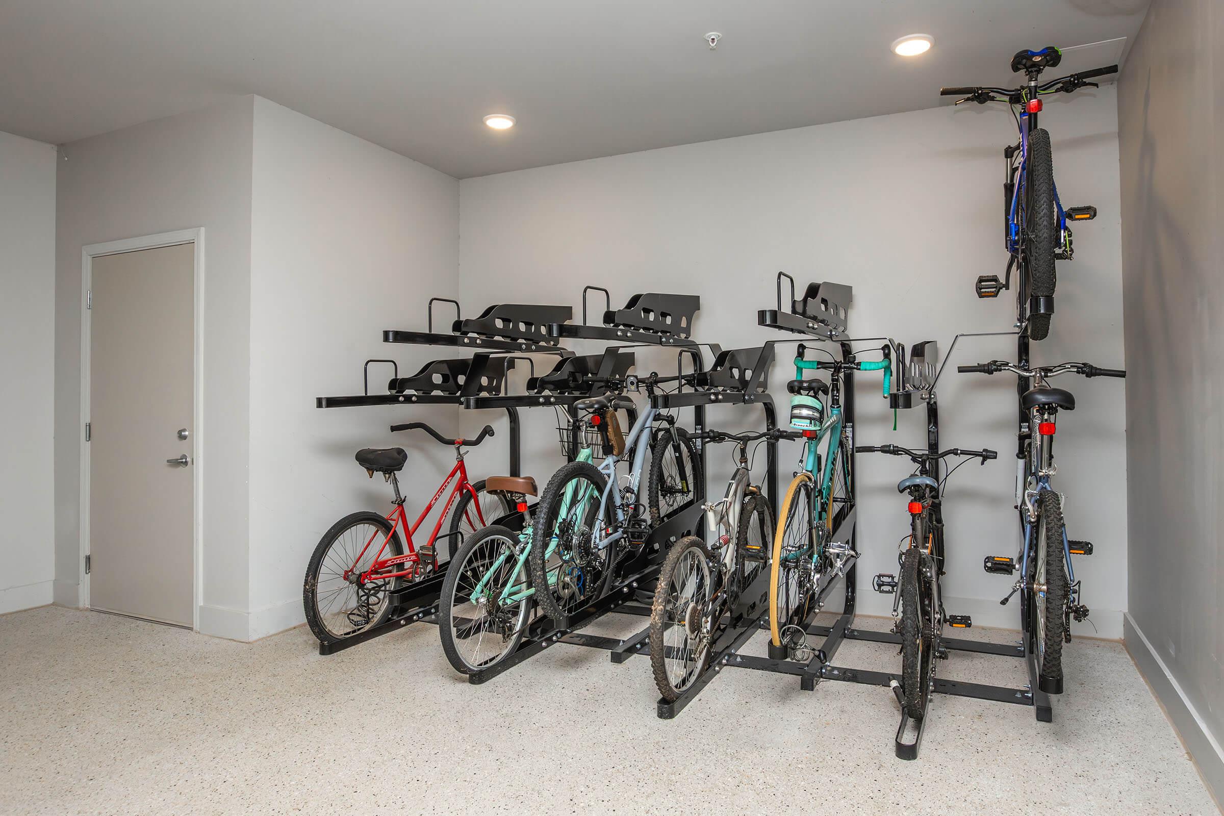 A well-organized indoor bike storage area featuring a variety of bicycles on racks. The space has a light gray wall, and the floor is made of a speckled material. There are two rows of bikes, including mountain and city bikes, in different colors and styles, neatly arranged for easy access.