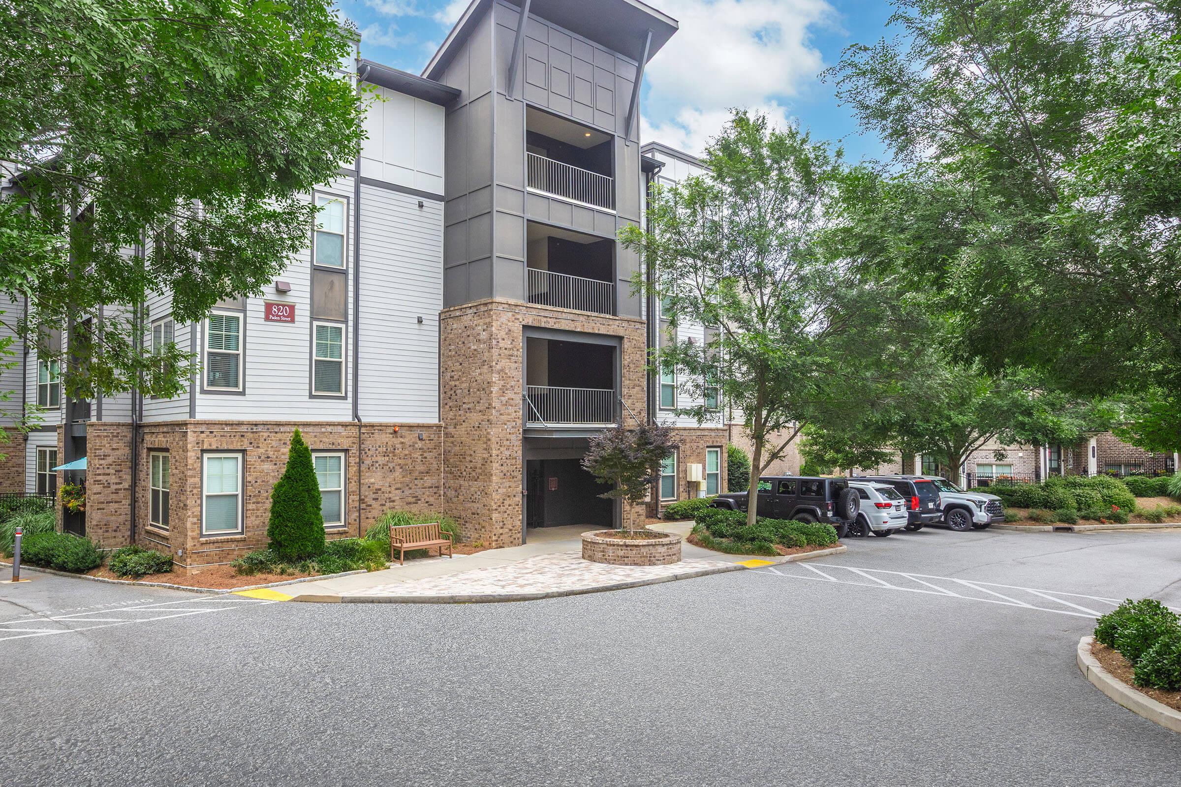 A multi-story residential building with a modern design, featuring a combination of stone and siding exterior. There are trees and landscaping around the entrance, and a parking area with several cars. A bench is situated near the front, with a clear blue sky overhead.