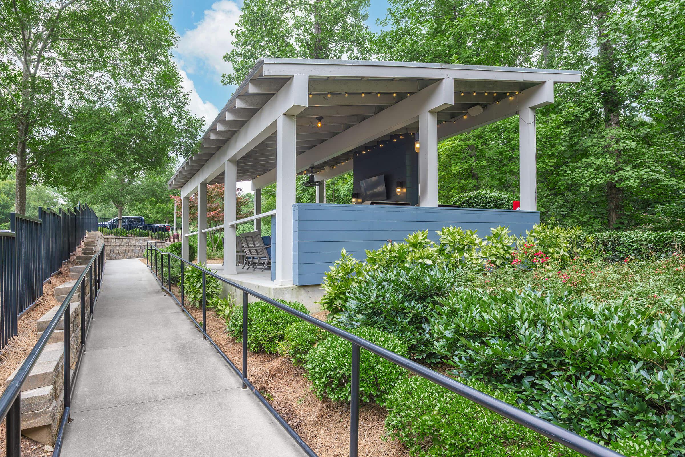 A covered outdoor seating area with a blue wooden structure is surrounded by lush greenery. The path leading to it is lined with bushes, and there are stairs on the side. Soft lighting can be seen under the canopy, creating a welcoming atmosphere.
