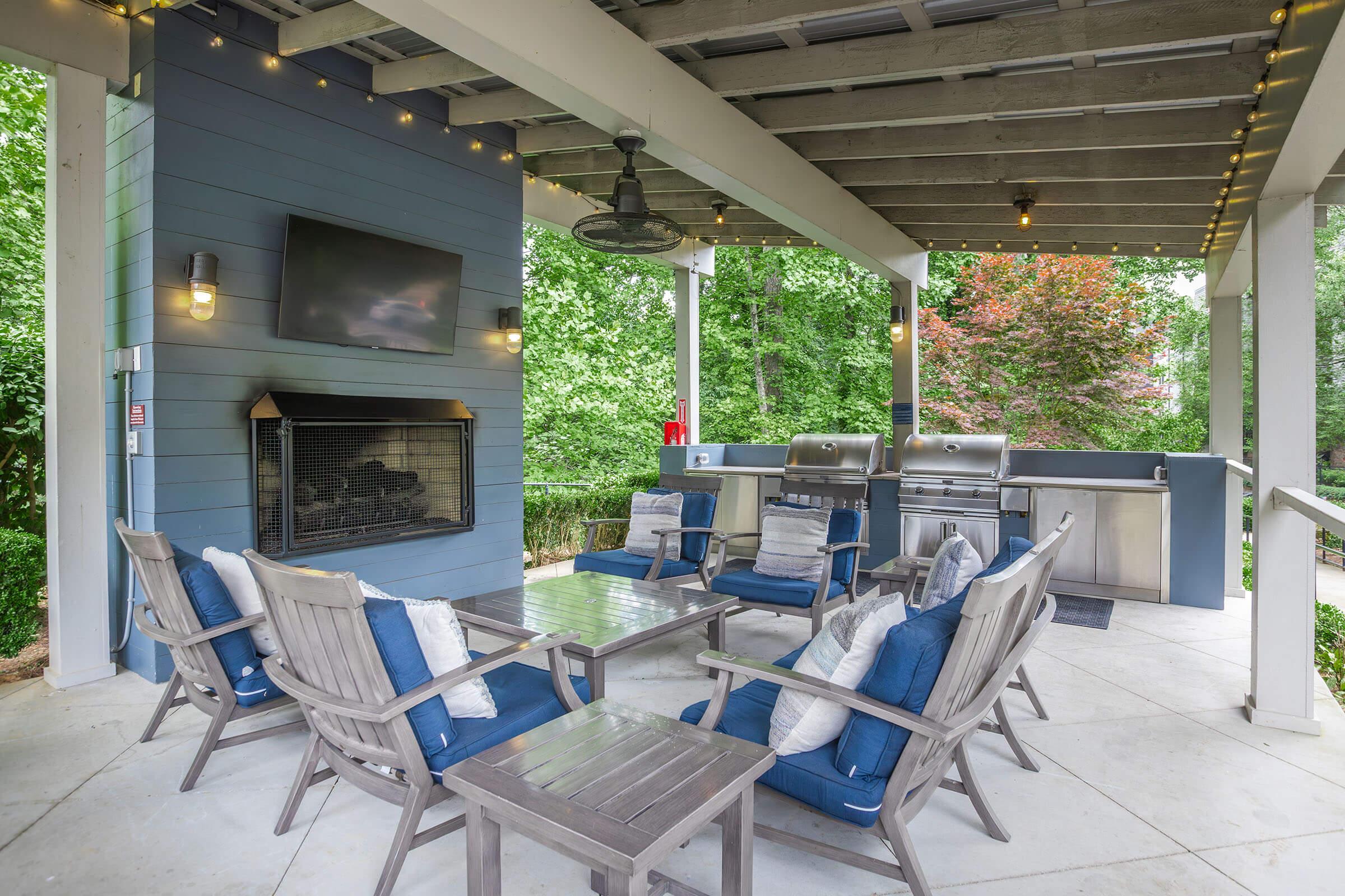 Outdoor patio space featuring comfortable seating with blue cushions, a modern outdoor fireplace, and a kitchen area with stainless steel grills. Green trees surround the space, creating a peaceful atmosphere. The ceiling is decorated with string lights for ambiance.
