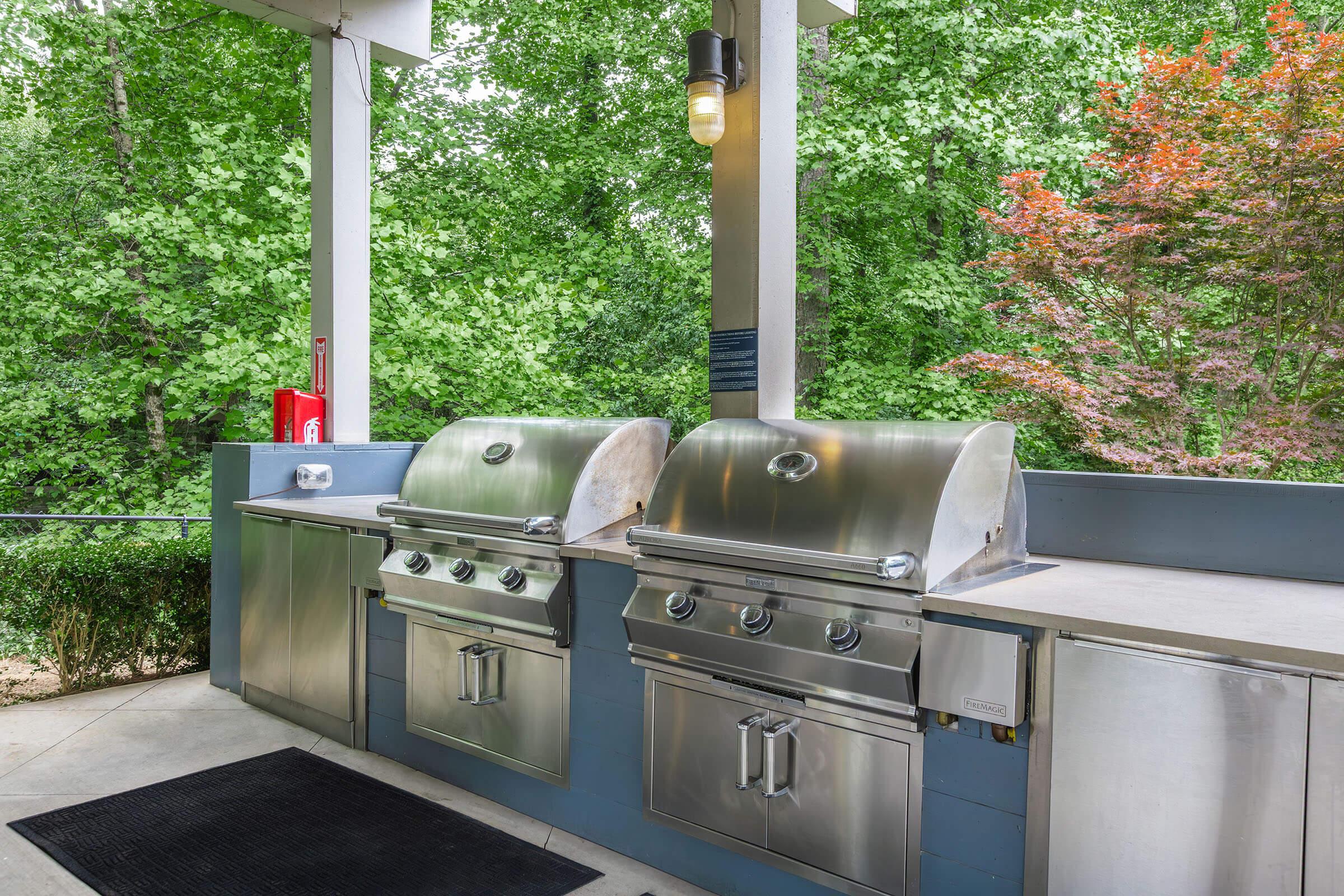 Outdoor cooking area featuring two stainless steel grills, a sink, and cabinets. The space is surrounded by lush greenery, with trees visible in the background. A light fixture hangs above the grill area, and there's a fire extinguisher mounted on the wall for safety.