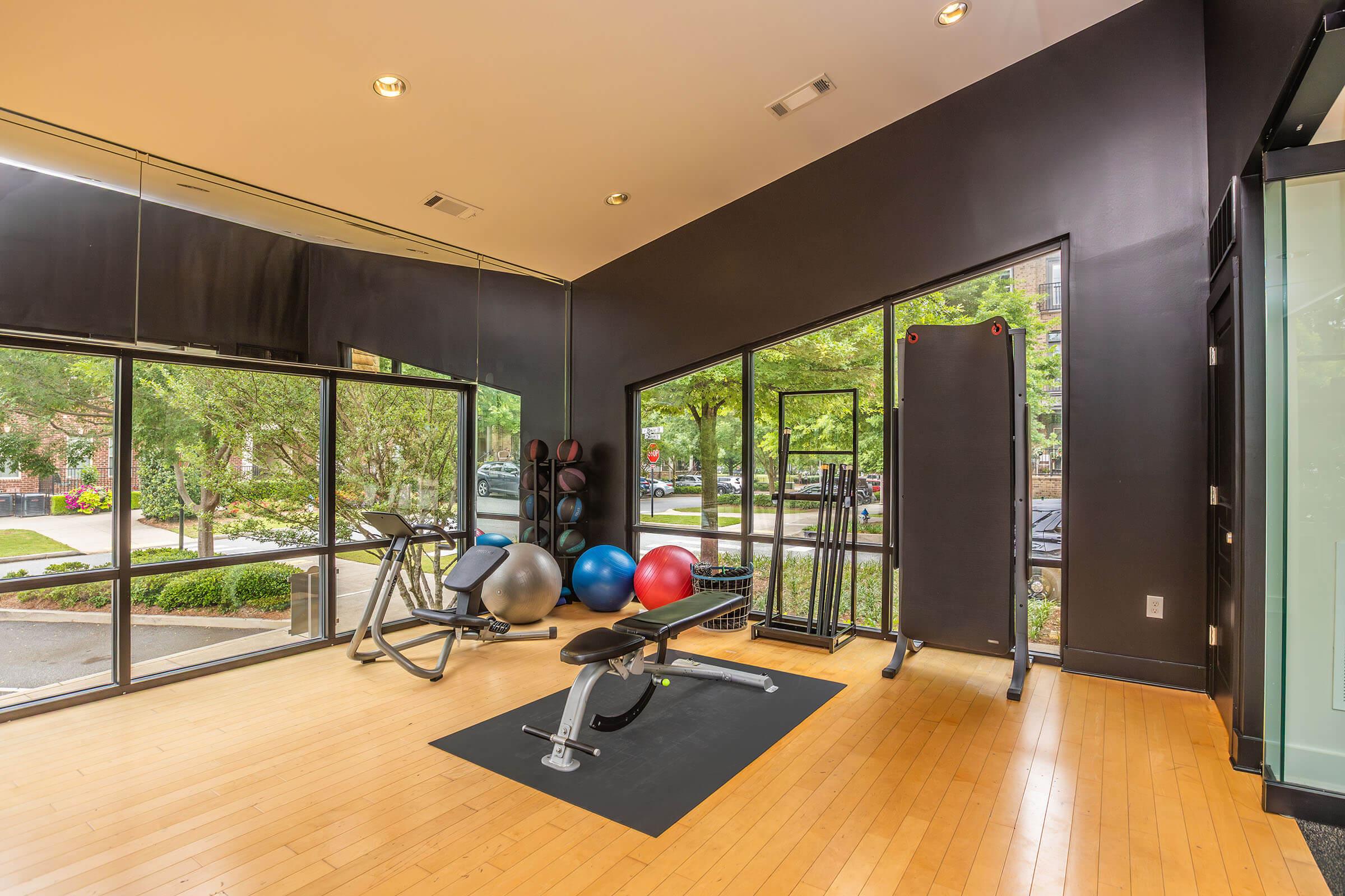 A modern gym interior featuring exercise equipment such as a bench press, free weights, and exercise balls. Large windows provide a view of the outdoors, with greenery visible outside. The flooring is wooden, and the walls are painted black, creating a contemporary aesthetic.
