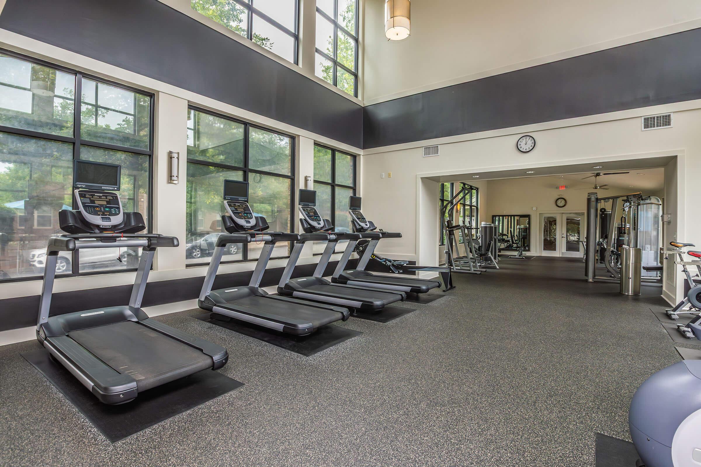 A modern gym interior featuring multiple treadmills lined up along a large window wall. The space has an open layout with natural light flooding in, rubber flooring, and additional gym equipment visible in the background. The atmosphere is bright and inviting for fitness enthusiasts.