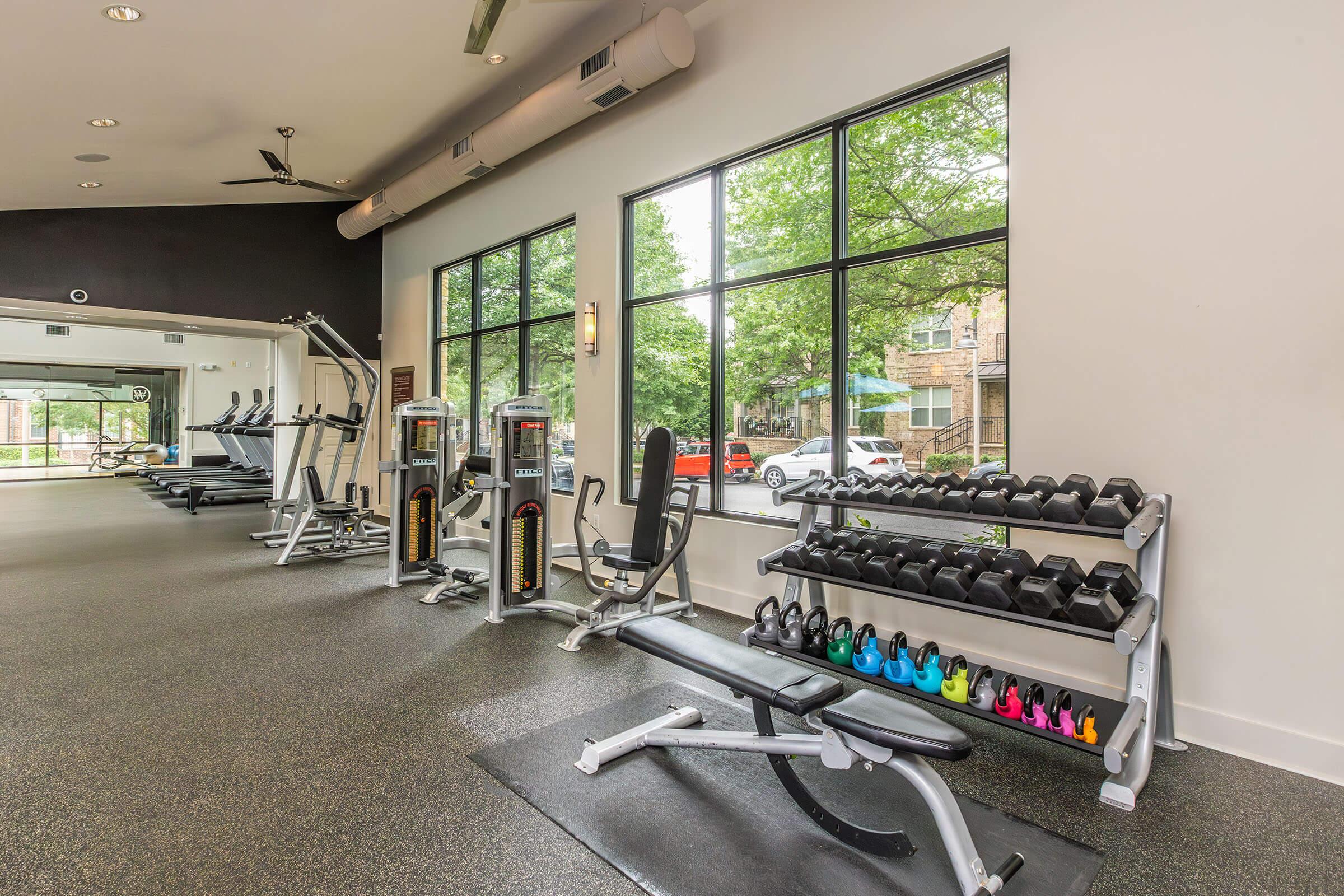 Interior of a modern gym featuring exercise equipment, including treadmills, weight machines, and a bench. Along one wall, there is a rack of colorful dumbbells. Large windows provide natural light and a view of greenery outside. The flooring is dark, and the space is well-organized.