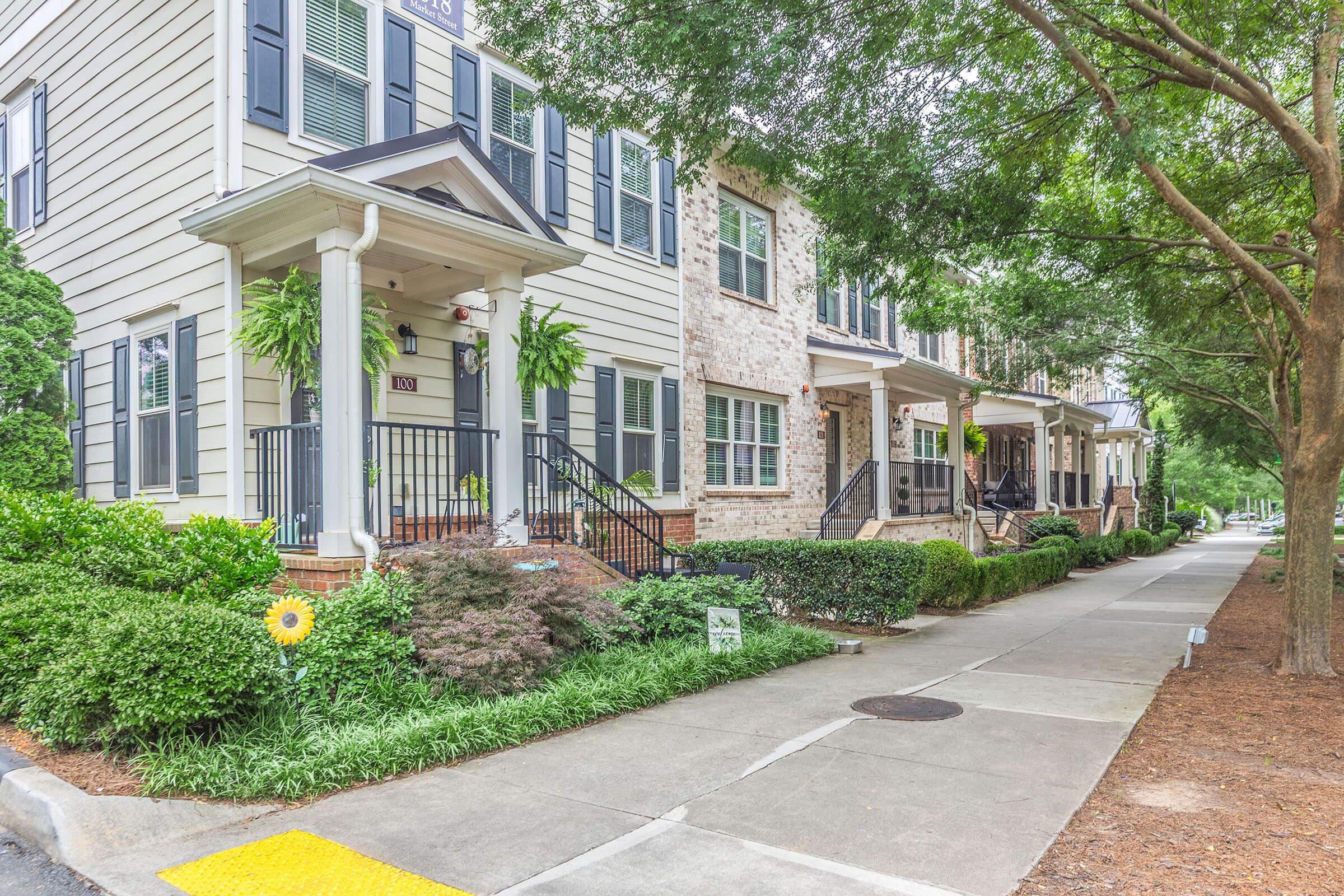 A row of charming two-story houses featuring a mix of siding and stone exteriors. Each home has a porch with decorative railings and plants. The sidewalk is lined with lush greenery and trees, creating a pleasant neighborhood atmosphere.