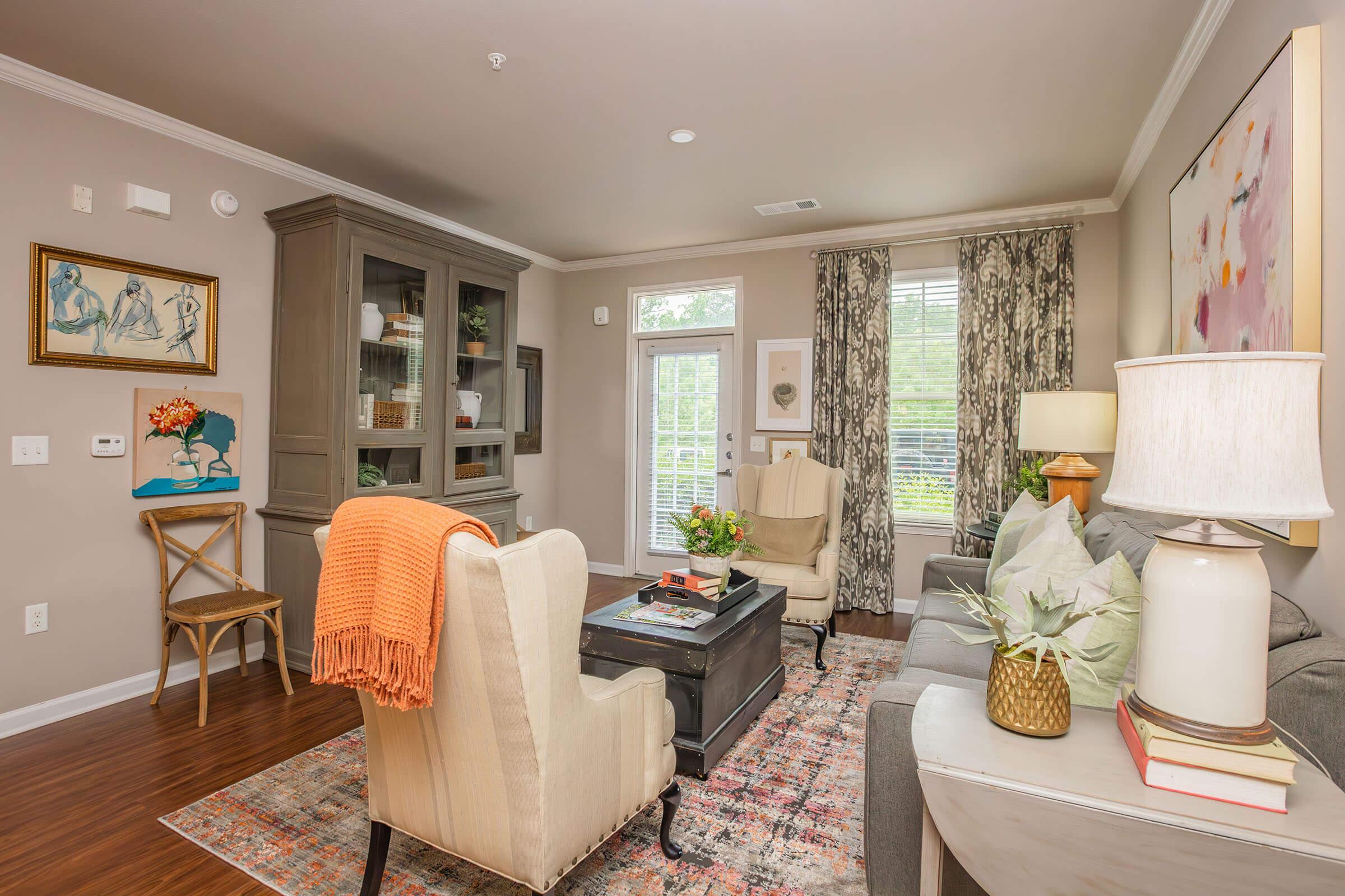 A cozy living room featuring a gray sofa, two armchairs, a decorative rug, and a coffee table. Natural light streams through large windows adorned with curtains. Artwork and plants add color to the space, while a bookshelf and modern lighting complete the inviting atmosphere.