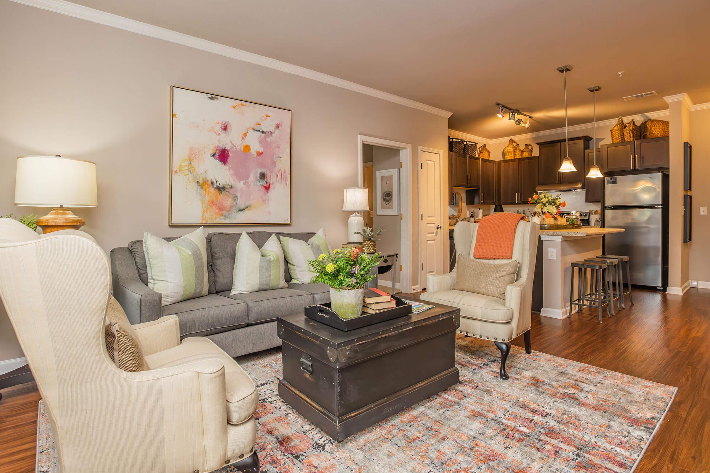 A stylish living room featuring a gray sofa with colorful pillows, two light armchairs, and a black coffee table. There's a vibrant abstract painting on the wall, hardwood flooring, and a well-lit kitchen area in the background with cabinets and decorative items.
