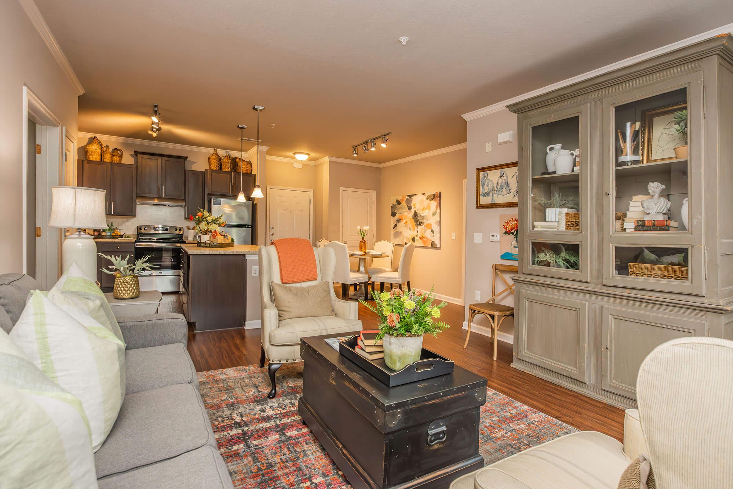 Cozy living room featuring a comfortable gray sofa, an accent chair with an orange throw, a dark coffee table, and a decorative cabinet with various knickknacks. The space has warm lighting, wooden flooring, and connects to a dining area with a table, creating an inviting atmosphere.