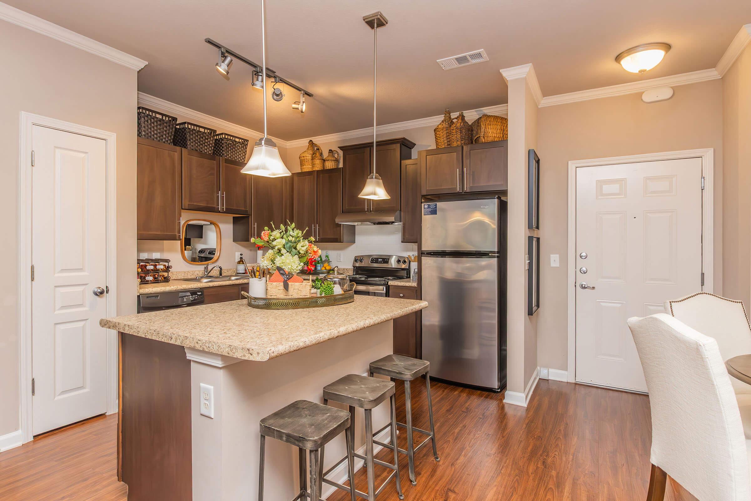 A modern kitchen featuring dark wood cabinetry, stainless steel appliances, and a granite island with bar stools. Decorative baskets line the shelves, and a vase of fresh flowers sits on the island. The space has warm lighting, an inviting atmosphere, and a door leading outside.