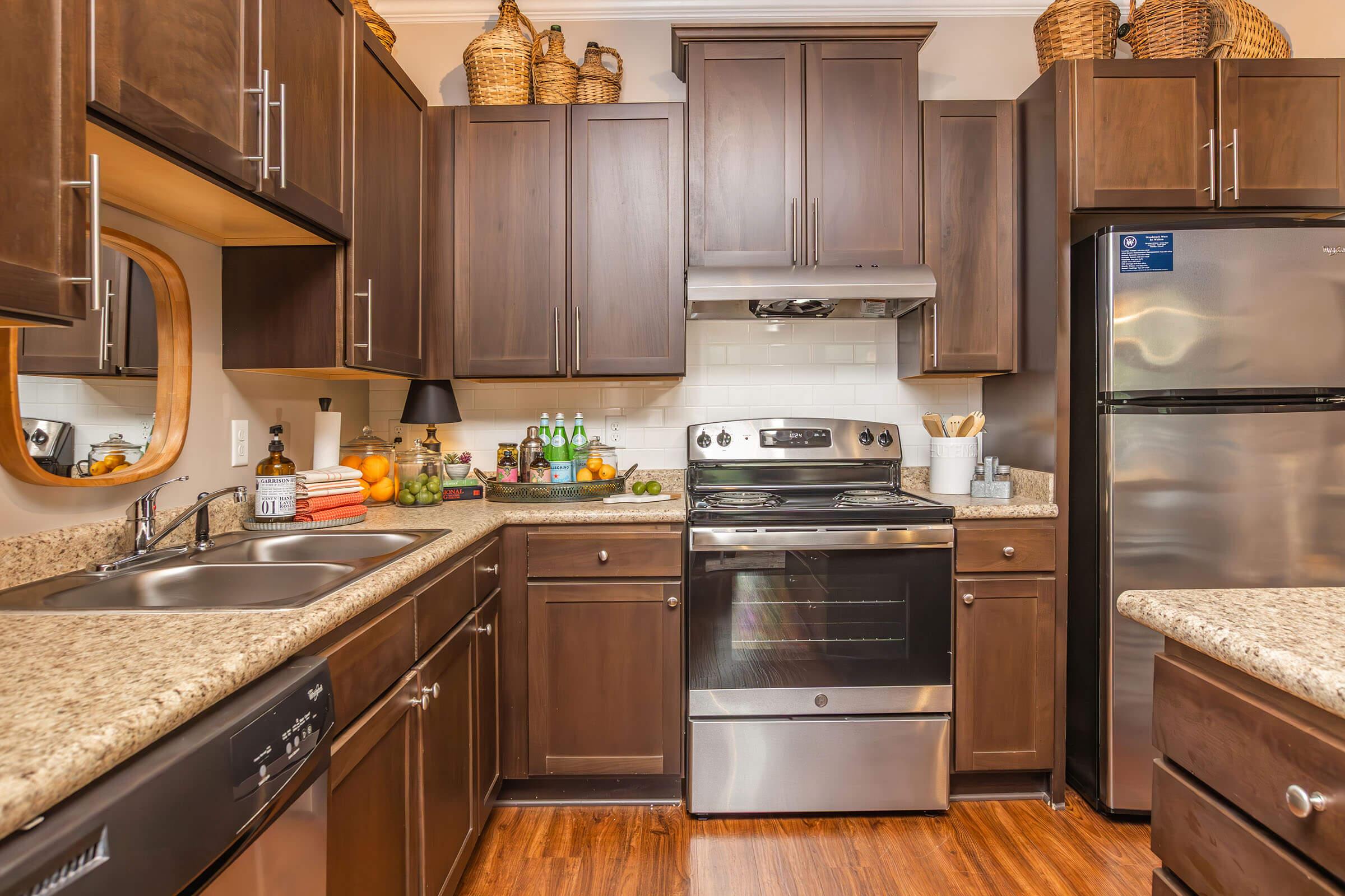A modern kitchen featuring dark wooden cabinets, a stainless steel stove and refrigerator, a granite countertop, and a sink. The space is decorated with baskets on top of the cabinets and various kitchen items, including green bottles and fresh fruits on the counter. Natural light illuminates the area.