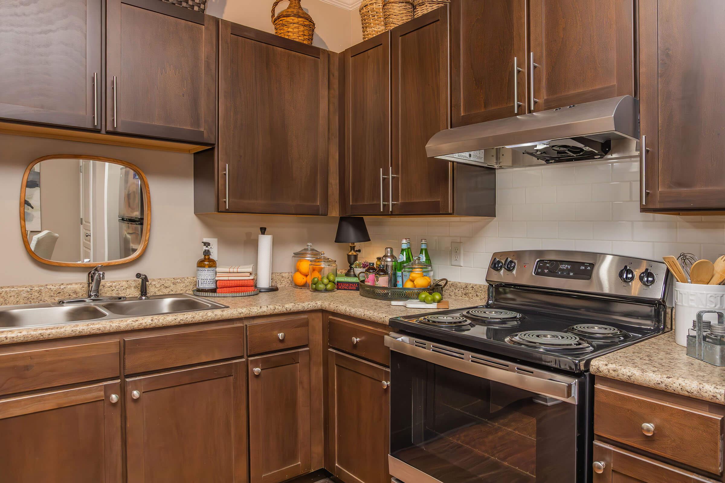 Modern kitchen featuring dark wood cabinets, a stainless steel stove, and a double sink. The countertop is made of granite and includes various kitchen accessories like bottles and fruits. A mirror is mounted on the wall, and shelves hold decorative baskets and beverages. Soft lighting adds warmth to the space.