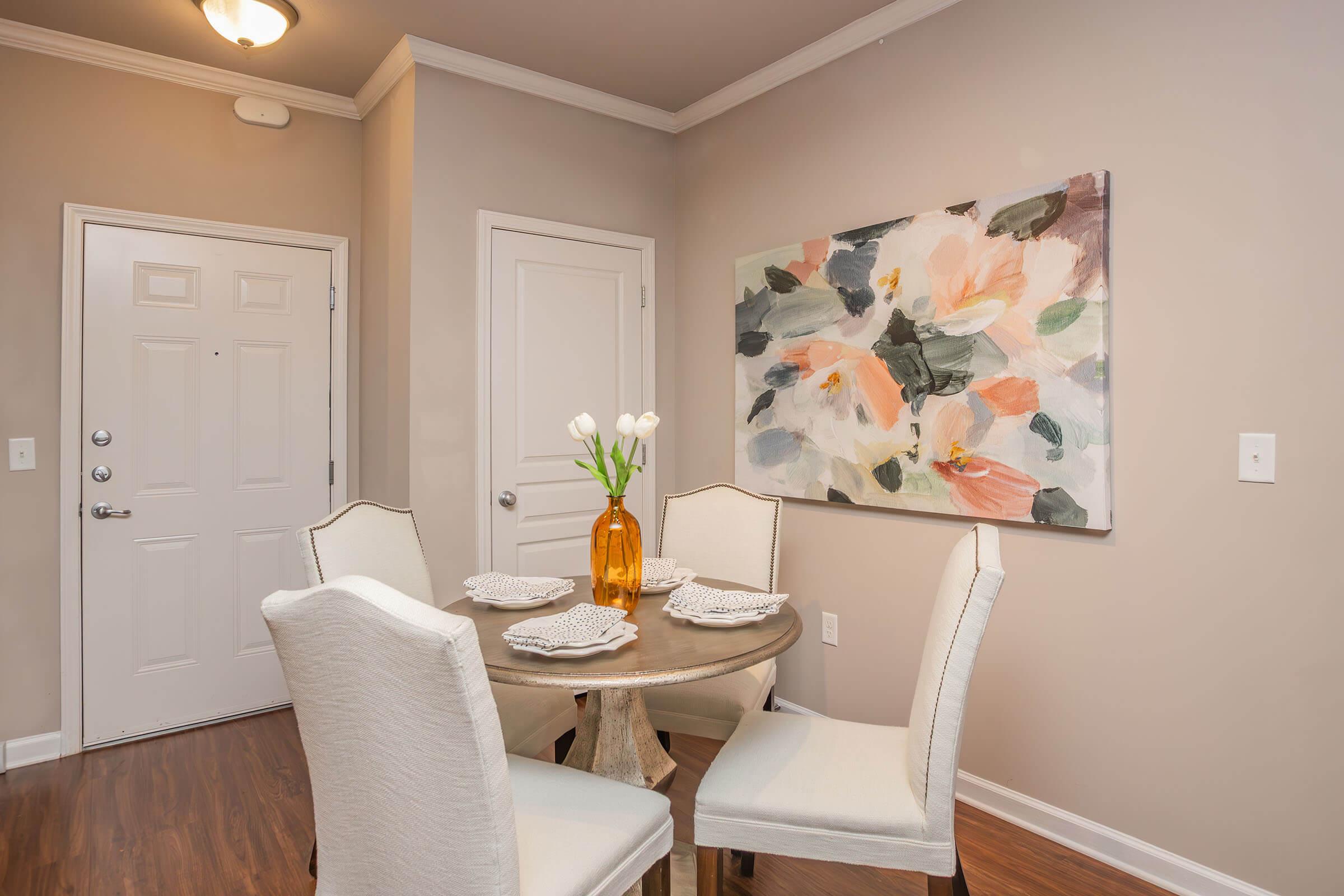 A cozy dining area featuring a round wooden table set with four white chairs and decorative plates. In the background, a light-colored door and wall complement a large floral painting with soft pastel colors. A vase with fresh white tulips sits at the center of the table, adding a touch of elegance.