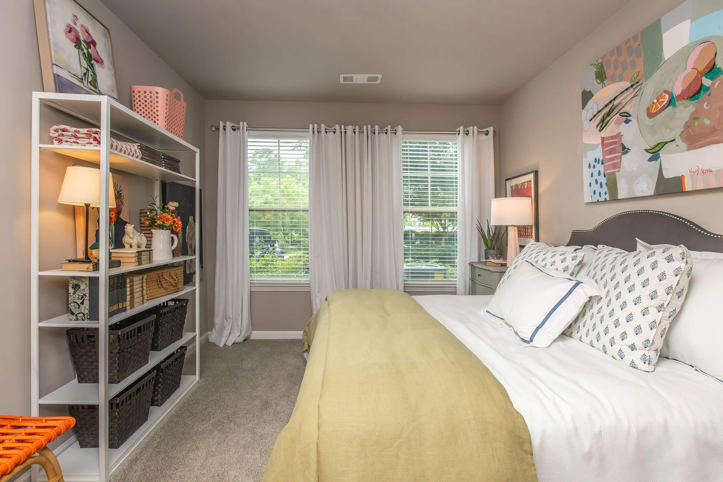 A cozy bedroom featuring a neatly made bed with decorative pillows, a light green throw blanket, a stylish nightstand with a lamp, and a shelf adorned with plants and decorative items. Natural light streams through a window covered with sheer curtains, enhancing the warm ambiance of the space.