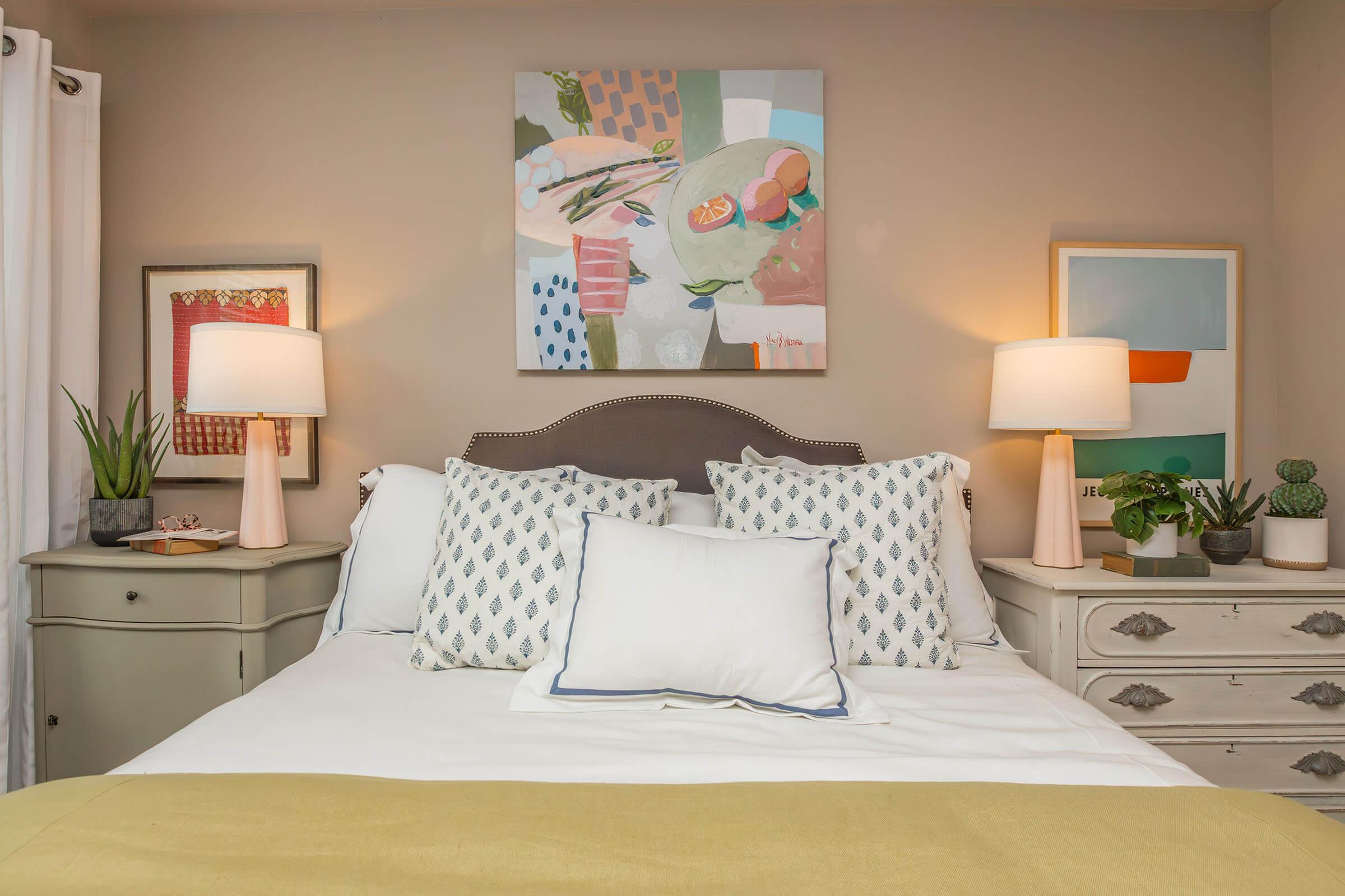 A cozy bedroom featuring a neatly made bed with decorative pillows, two bedside lamps with pink bases, and a colorful abstract painting above the bed. There are potted plants and framed artwork on the walls, creating a warm and inviting atmosphere. The color palette includes soft pastels and neutral tones.