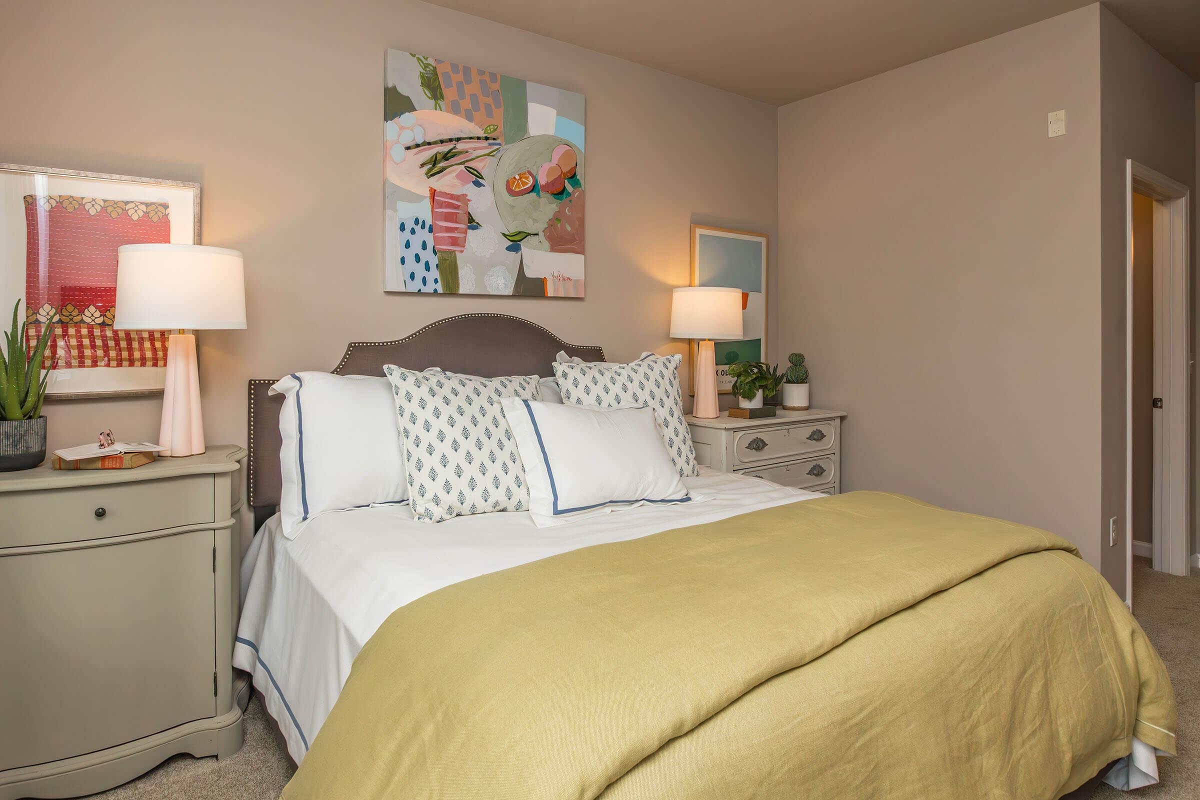 A cozy bedroom featuring a queen-sized bed with a light yellow blanket, decorative pillows, and a modern headboard. The room includes two bedside lamps, a stylish nightstand, and a dresser. Artwork hangs on the wall, and there's a plant and mirror, enhancing the inviting atmosphere. Soft, neutral colors create a tranquil ambiance.