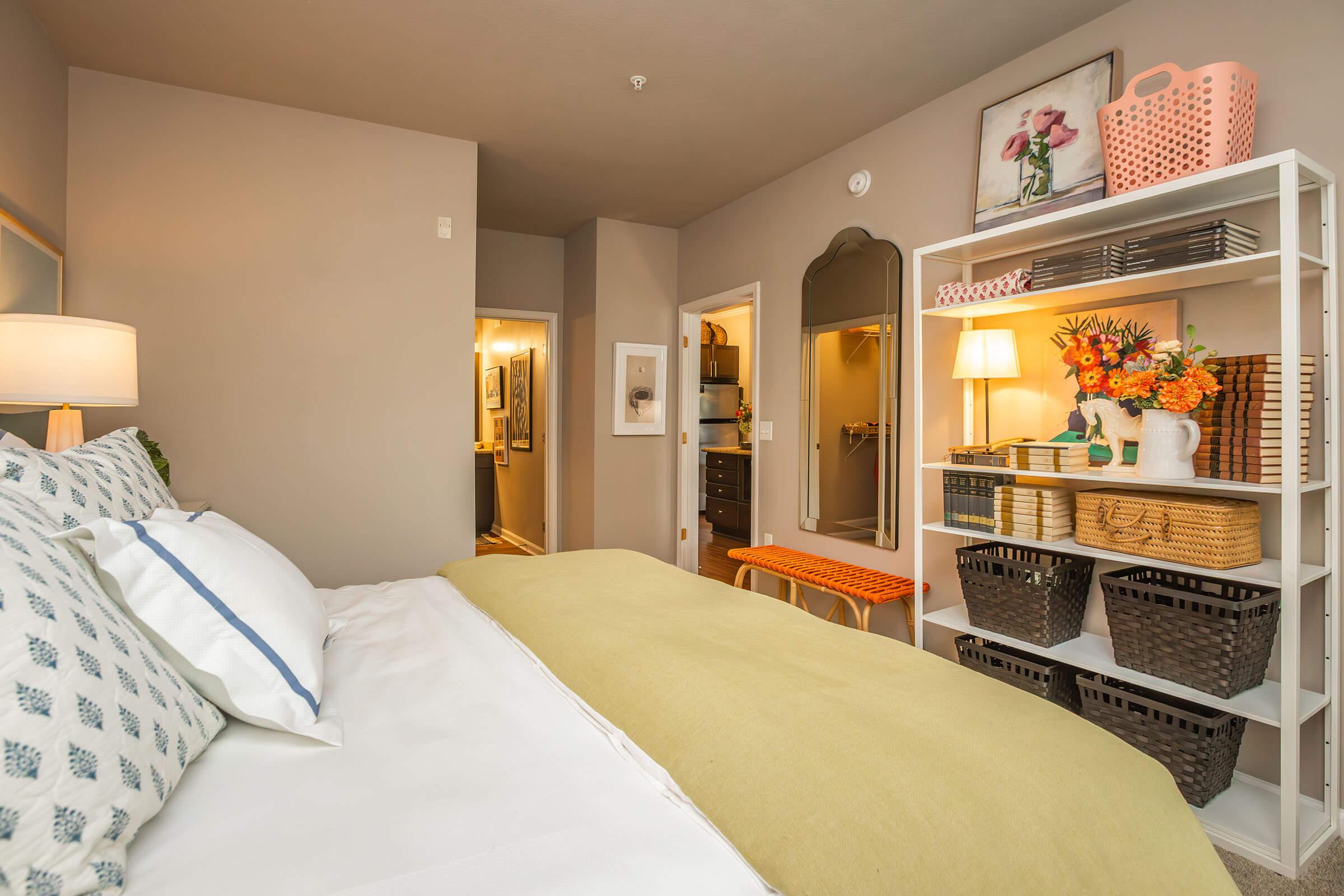 A cozy bedroom featuring a neatly made bed with a light green throw blanket, decorative pillows, and a bedside lamp. A white shelving unit displays baskets and flowers. The room includes a doorway leading to a bathroom and another door leading to a closet, with soft, neutral wall colors and warm lighting.