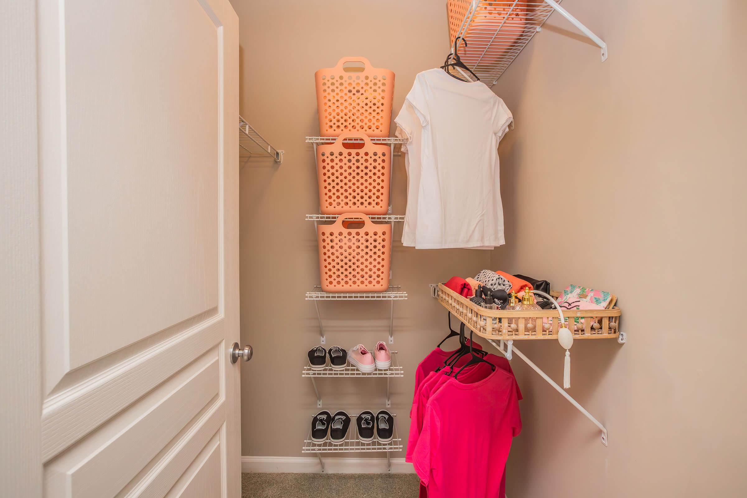 A neatly organized closet with light beige walls. It features three pink storage baskets on a shelf, a white shirt hanging, and a collection of shoes on a lower rack. On the right side, there are two pink shirts hanging from a hook, along with small items displayed in a woven basket on a shelf.