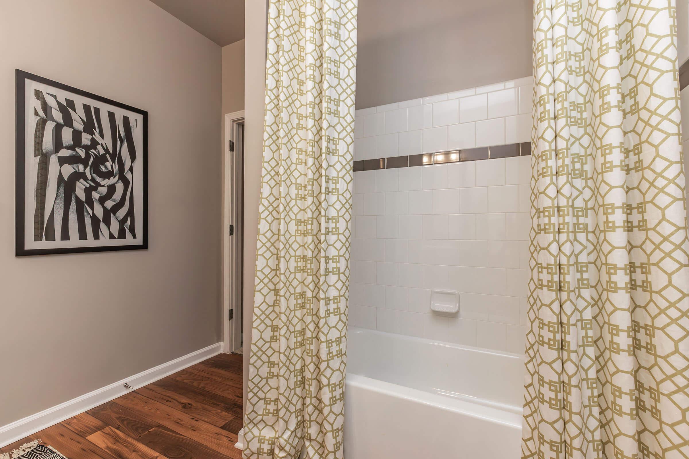 A bathroom featuring a white bathtub with a shower curtain, geometric patterned curtains in green and white, a decorative black and white abstract art piece on the wall, and wooden flooring. Natural light illuminates the space, creating a clean and modern aesthetic.